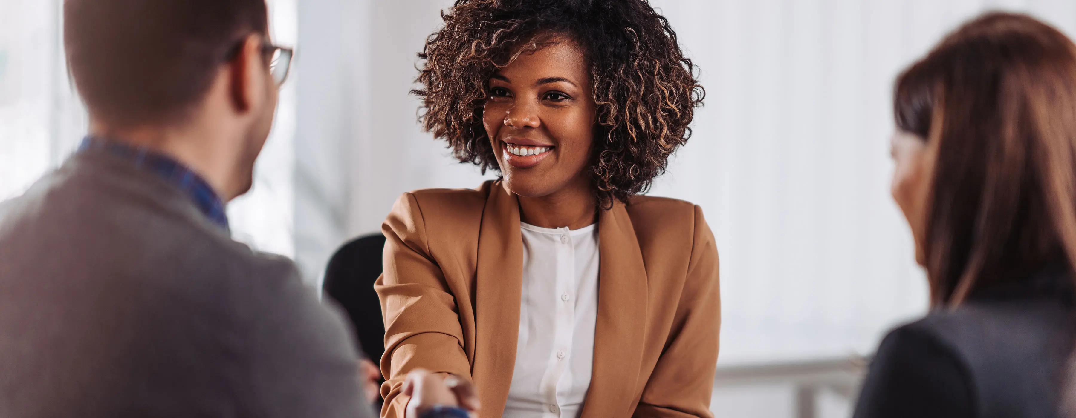 Foto de um mulher jovem sorridente apertando as mãos de uma outra pessoa a sua frente, como cumprimento e acordo de algum contrato fechado entre as partes e uma outra pessoa a frente observando como testemunha.