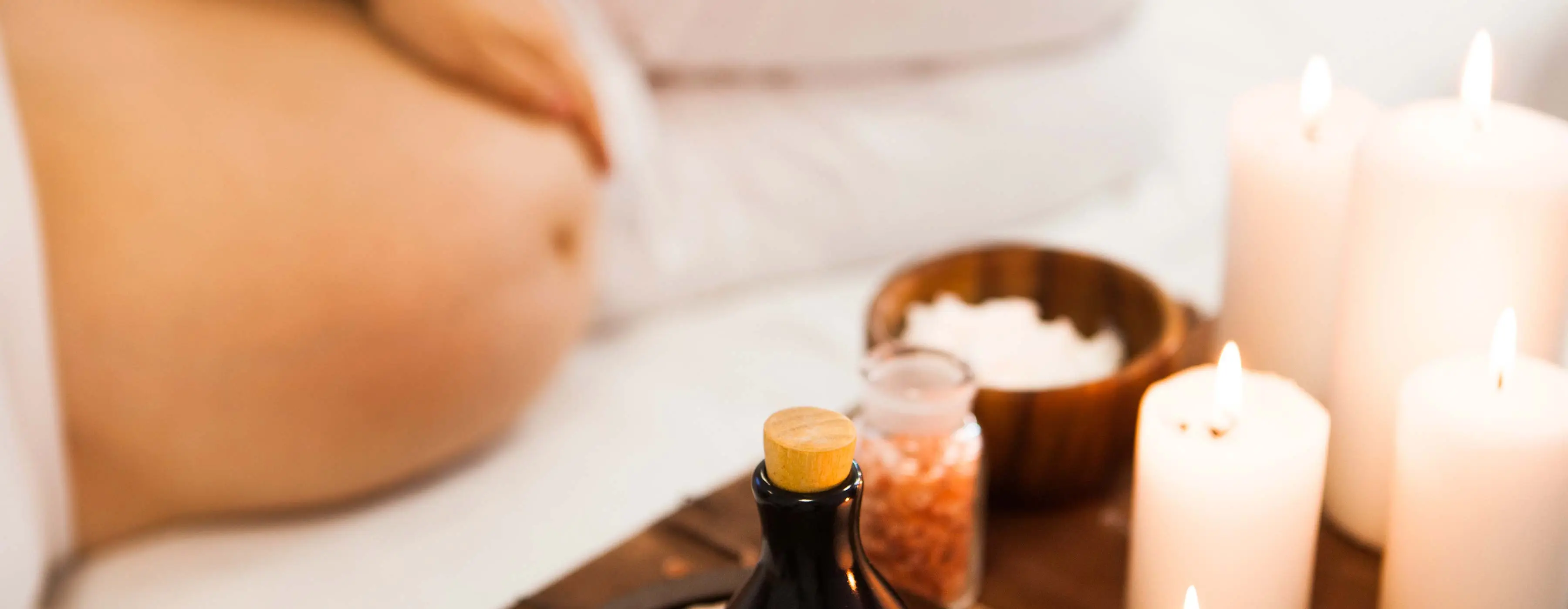 Foto em close de velas acesas e sais de banho. Ao fundo, gestante deitada com a mão na barriga.