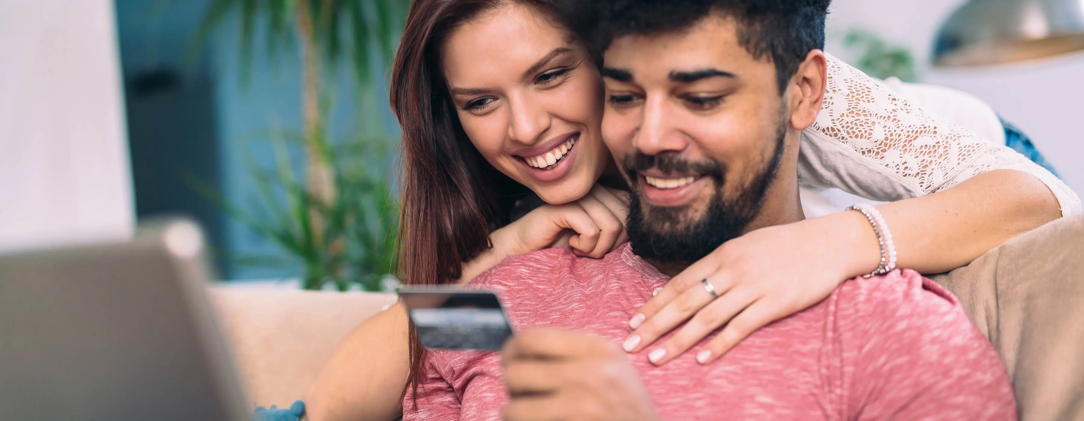Foto em close de um homem e uma mulher jovem sorridentes verificando um cartão de crédito e fazendo compras on line pelo notebook.