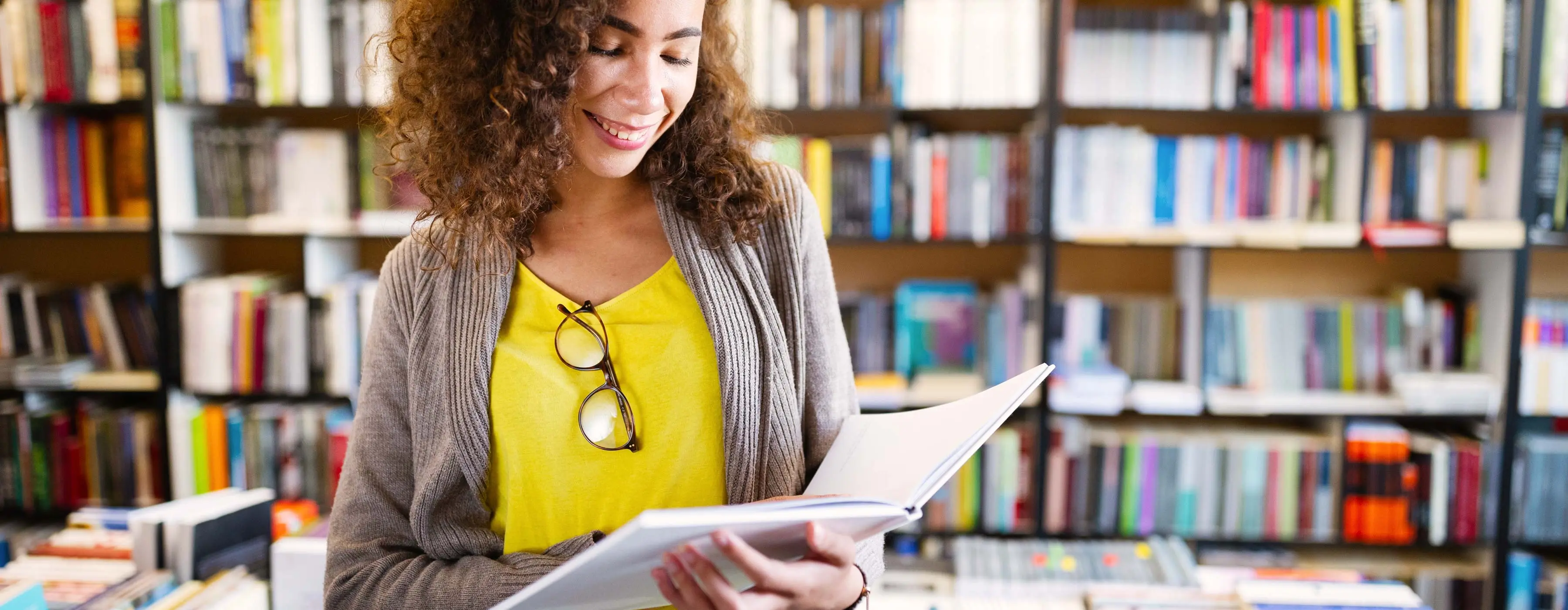 Foto de uma mulher jovem sorrindo, fazendo pesquisa e verificando um livro em uma biblioteca.