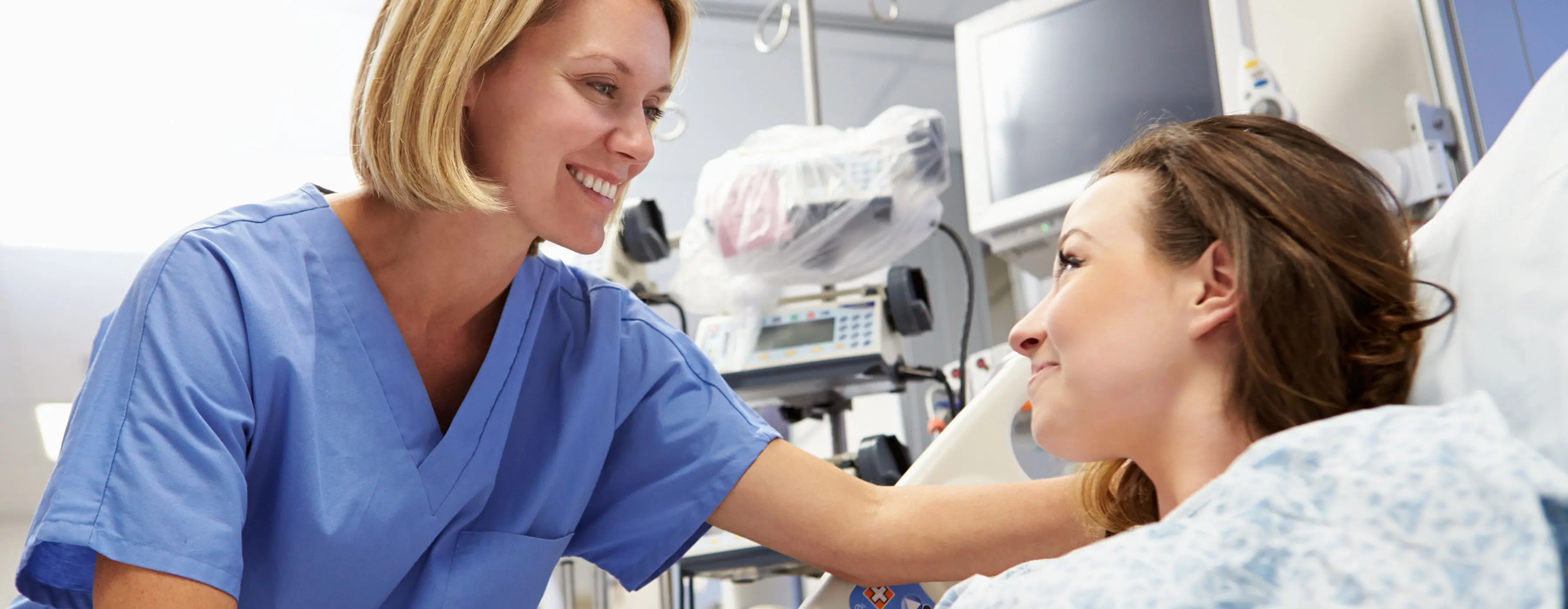 Foto de uma profissional da saúde, em quarto de hospital, com equipamentos ao fundo, sorrindo e olhando para paciente deitada na cama.