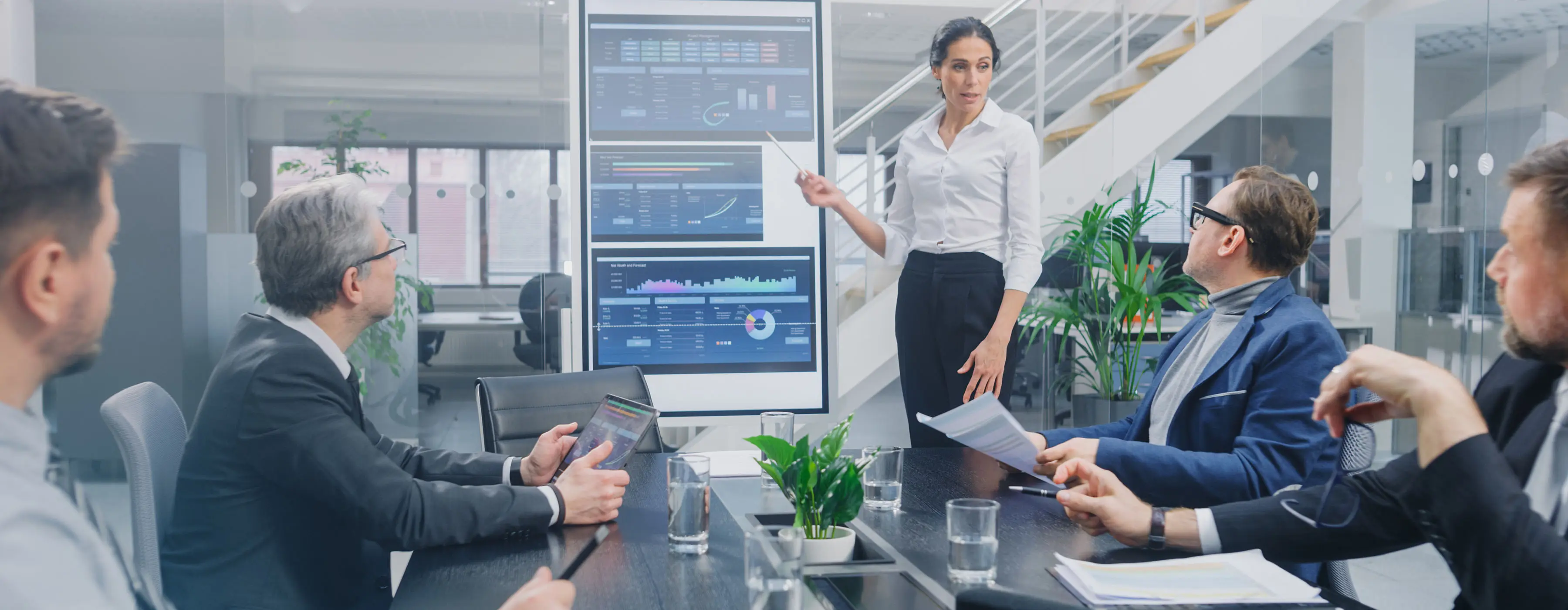 Foto de um grupo em uma sala de reuniões corporativas, algumas pessoas sentadas prestando atenção na apresentação de uma mulher em pé apontando um quadro interativo digital com gráficos.