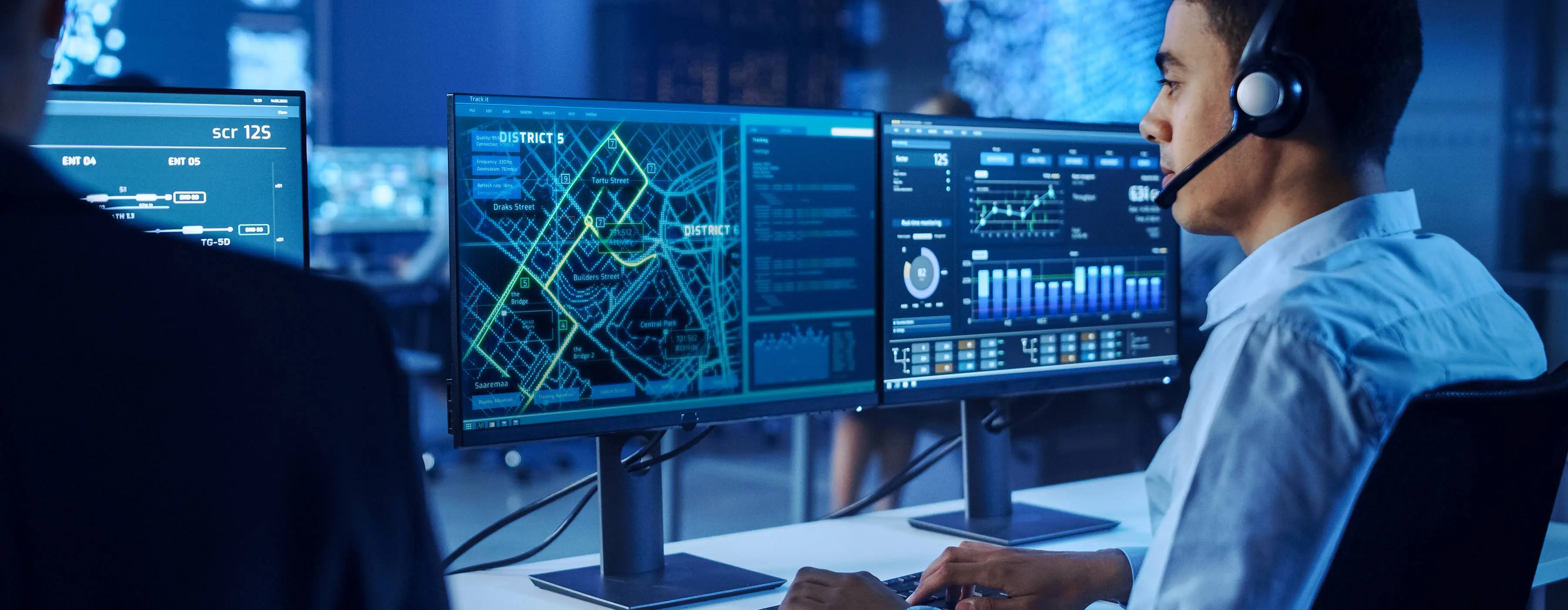 Foto de um homem jovem, em uma central de processamento de informações, observando imagens de mapas em dois monitores de computador.