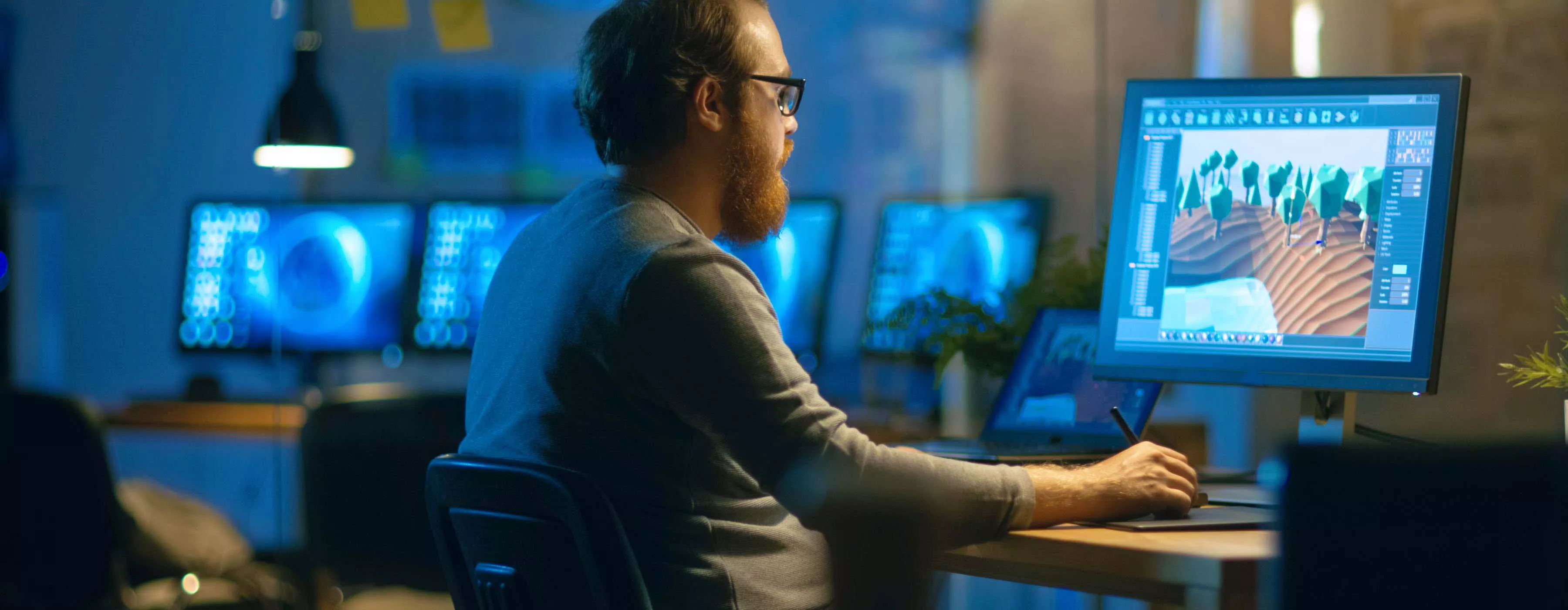Foto de um homem trabalhando sentado em um escritório, desenvolvendo um projeto gráfico em seu computador utilizando dois monitores.