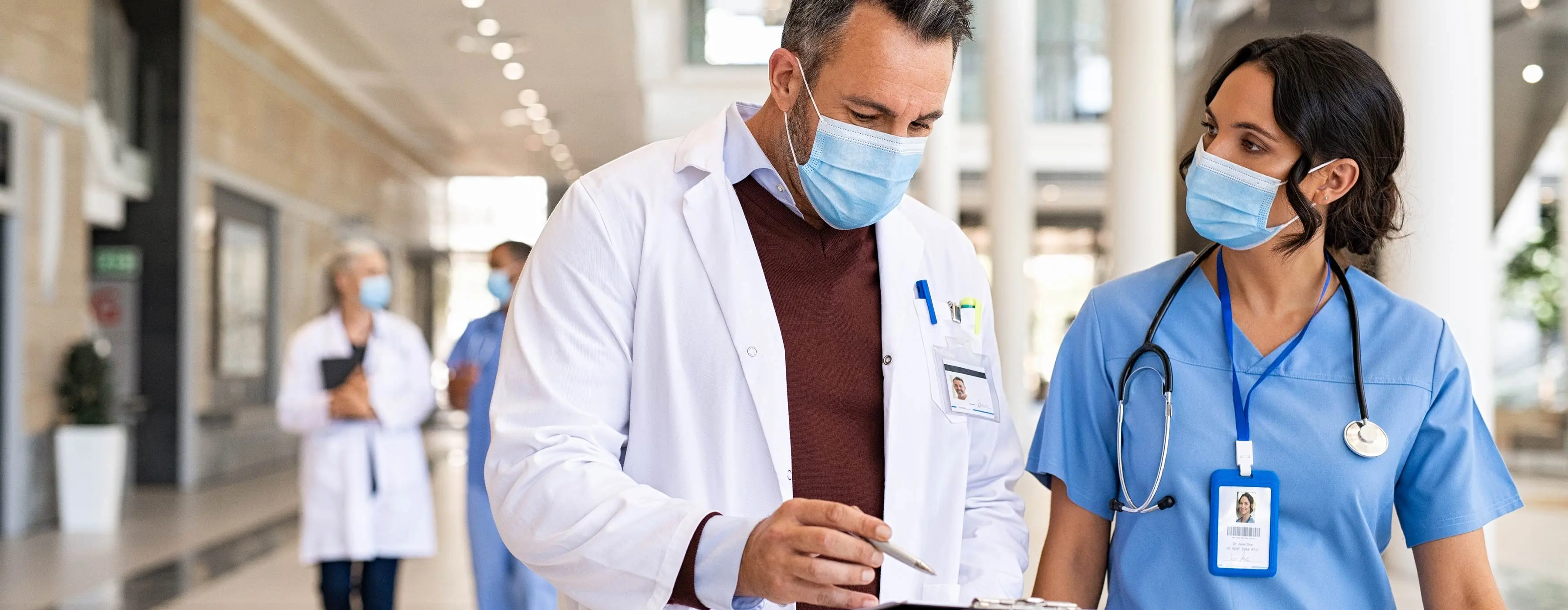 Foto de dois profissionais da saúde, em corredor de hospital, conversando e analisando dados de uma prancheta.