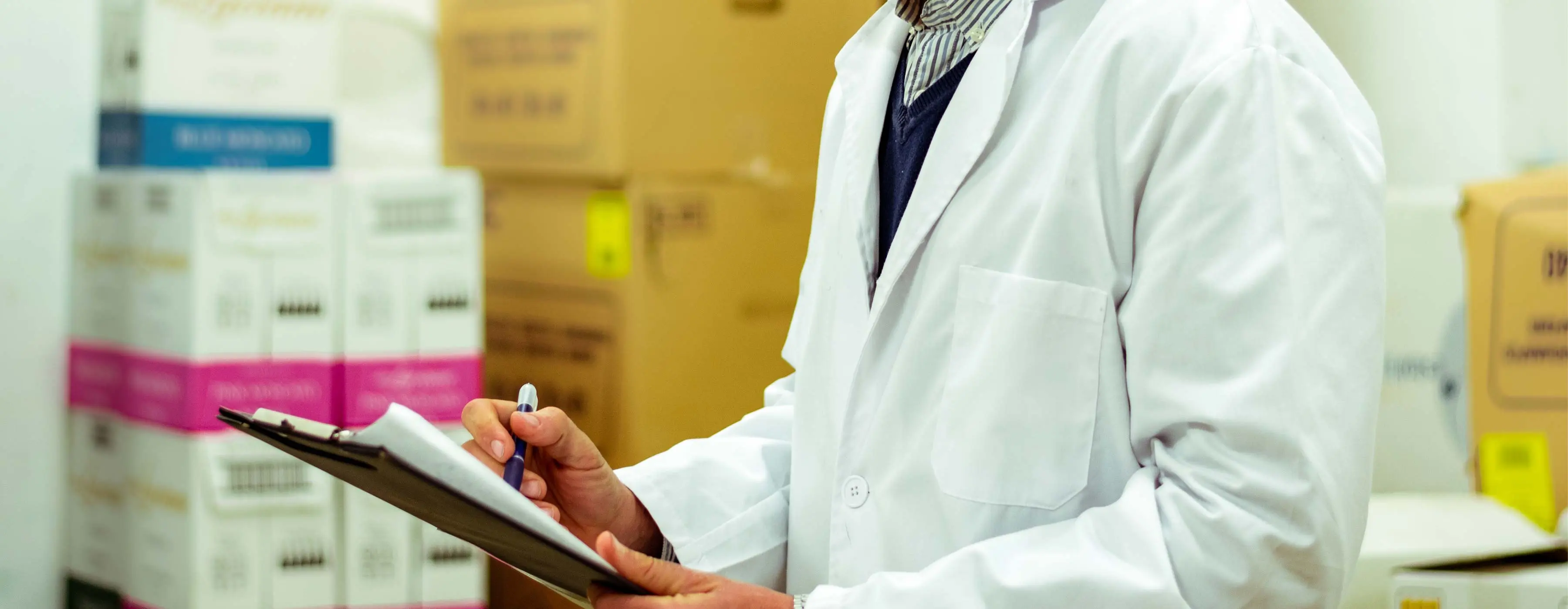 Foto em close das mãos de um profissional de saúde, no estoque de medicamentos, verificando documento em prancheta.