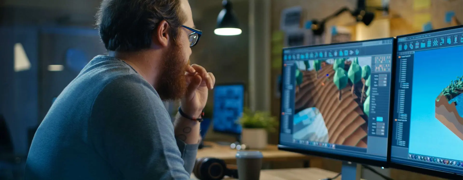 Foto de perfil com homem de óculos e barba ruiva em frente ao computador com dois monitores utilizando softwares para criação de jogos. Sozinho, ele está em um escritório com luz baixa, café e fone sobre a mesa