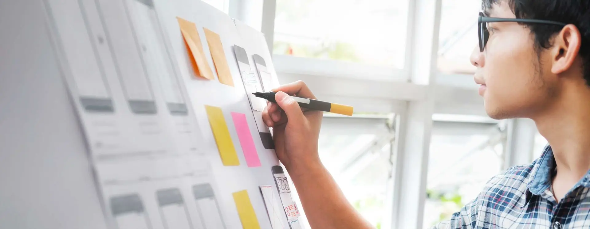 Foto de jovem de óculos utilizando a mão direita para escrever nos postit's do flipchart com canetão preto