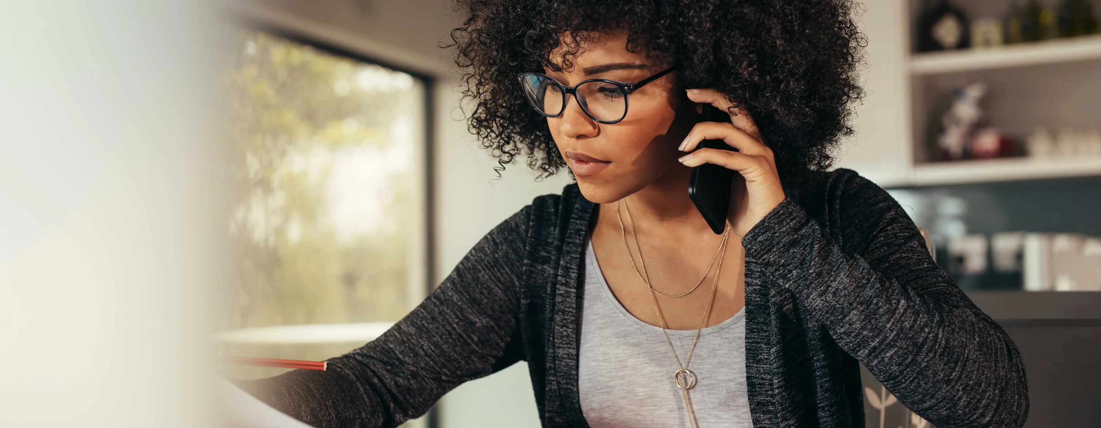Foto de uma mulher sentada, conversando ao telefone e verificando ao mesmo tempo informações em um documento.