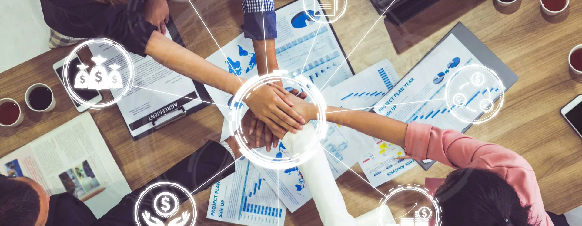 Foto de cima pra baixo em uma mesa de reuniões pegando mãos sobrepostas de profissionais demonstrando união de grupo, com cafés e documentos espalhados. Sobre a imagem, uma montagem contendo ícones com cifrões representando lucro financeiro.