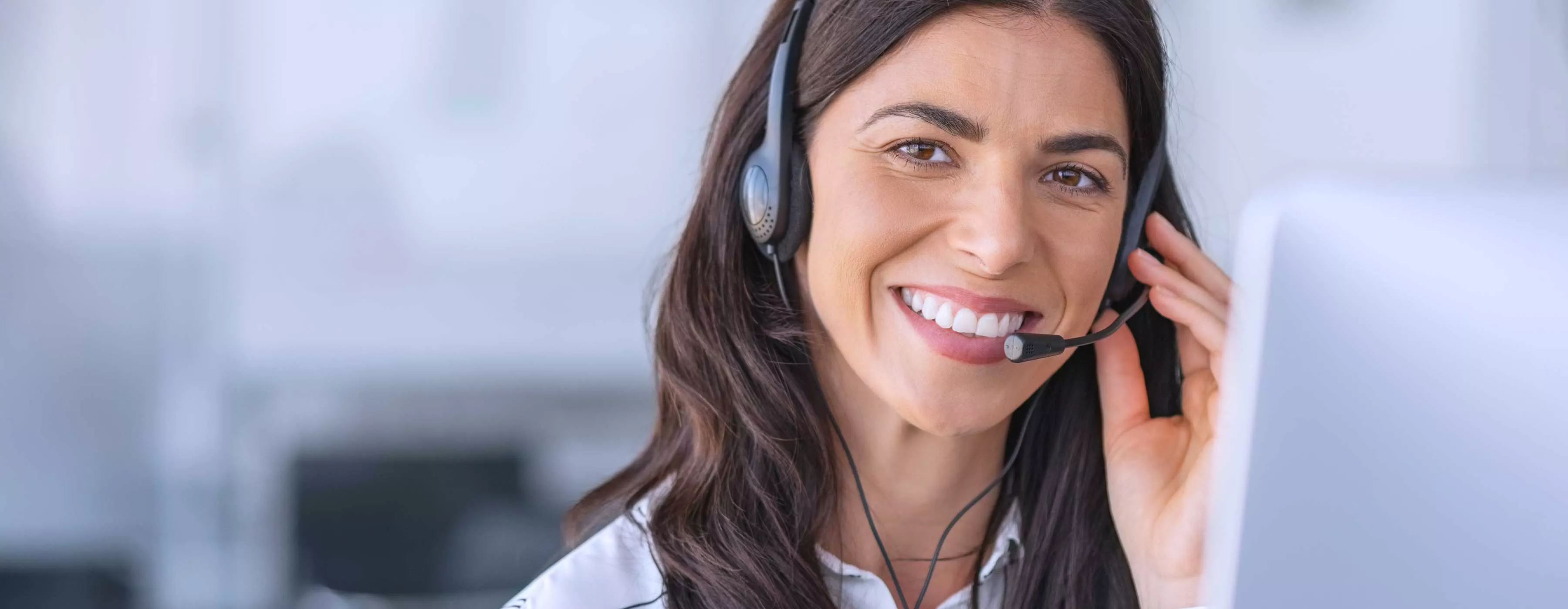 Foto de mulher sorrindo, sentada em frente a computador e com fone de ouvindo com microfone acoplado.
