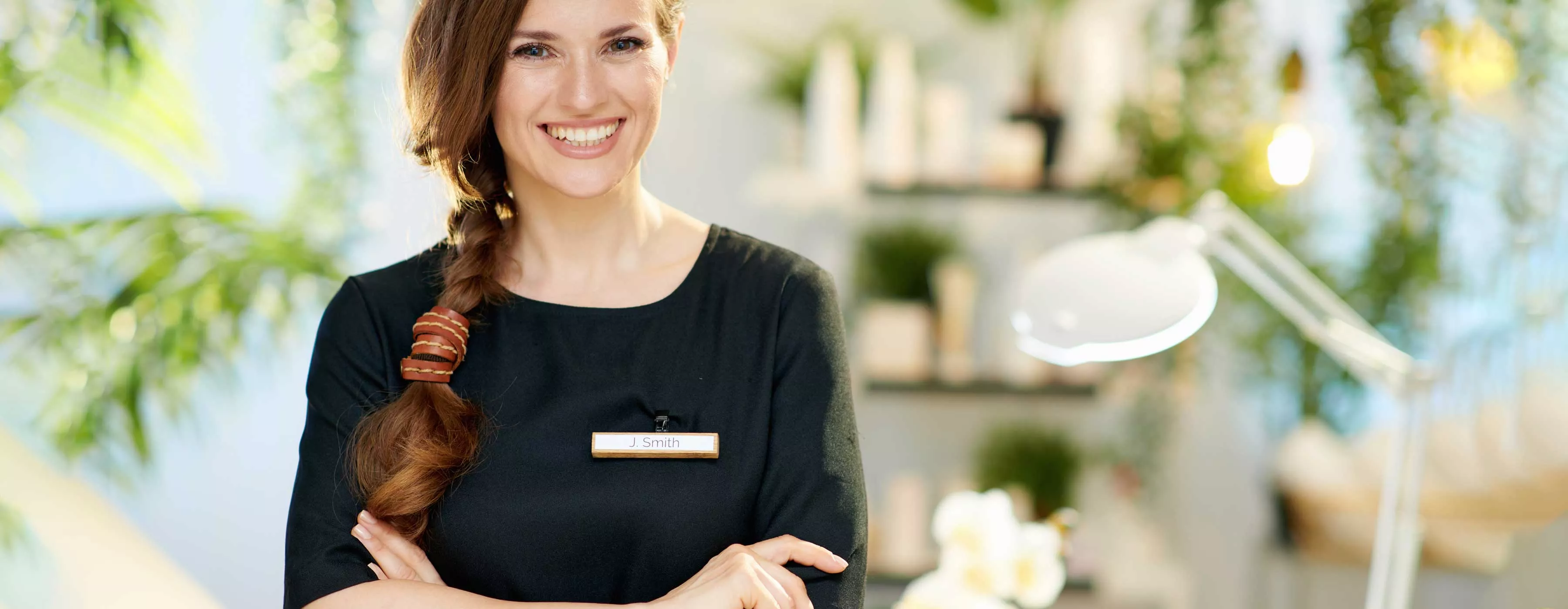 Foto de uma mulher sorrindo de braços cruzados com uniforme em ambiente de spa.
