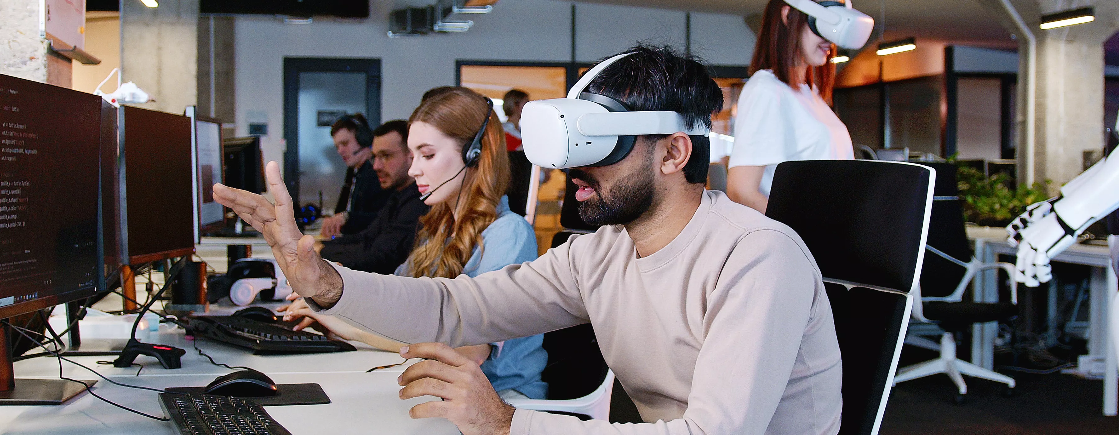 Foto de homem utilizando óculos de realidade virtual em frente a um computador. Ao fundo, na mesma sala, pessoas usam computadores também.