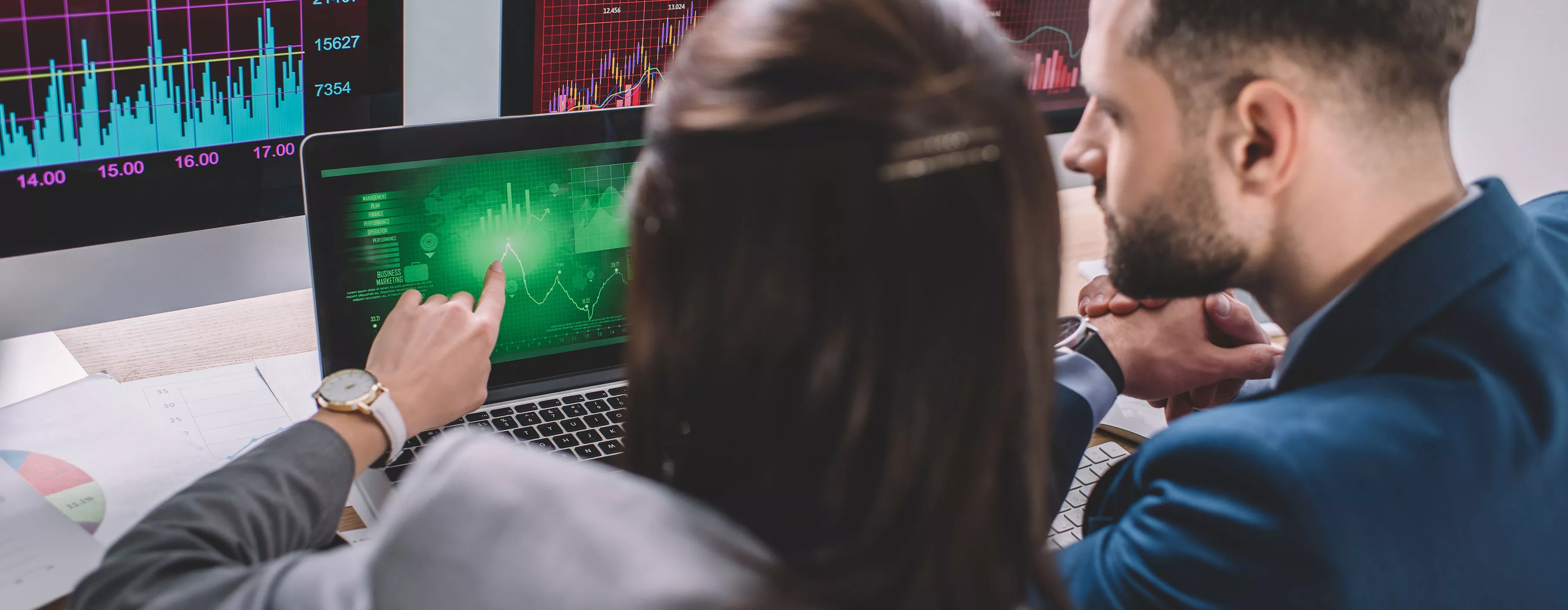 Foto de um homem e uma mulher mexendo em um computador com uma programação de software e ao fundo uma outra tela com um gráficos digitais.