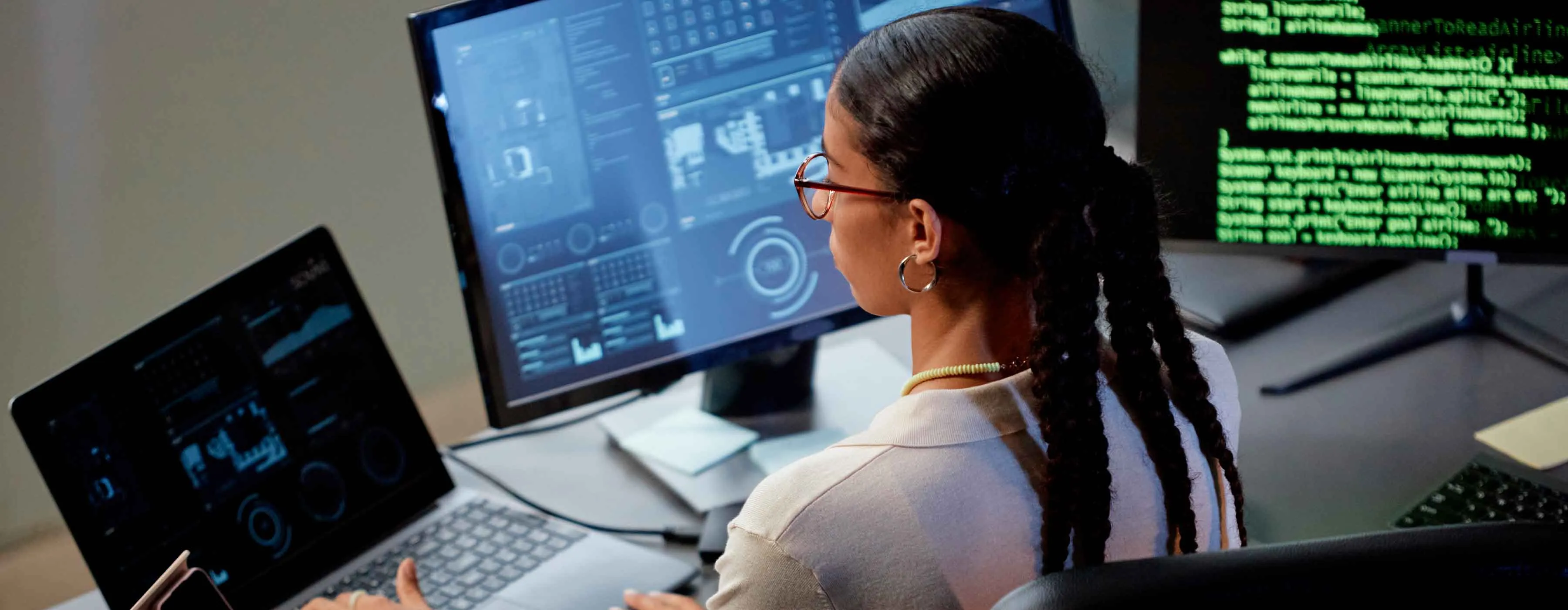 Mulher observa computador com 3 telas de dados e gráficos.