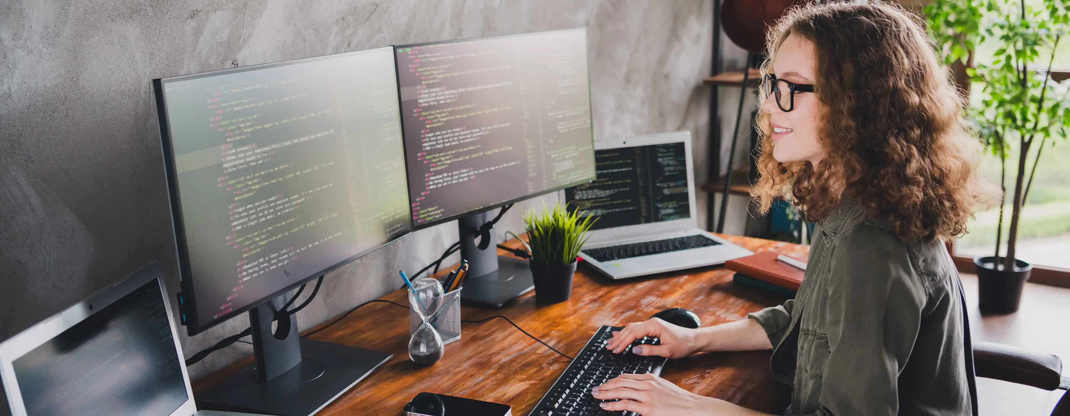 Mulher observa telas de computador exibindo código de programação.