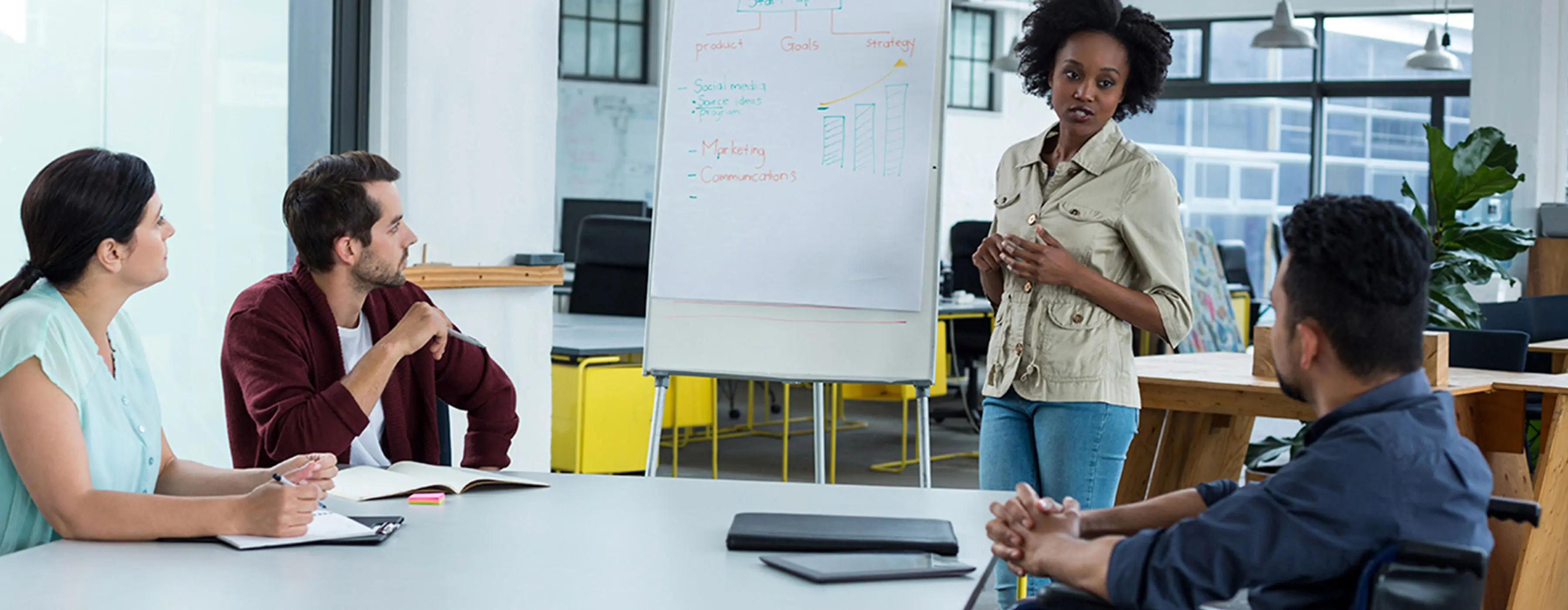 Foto de grupo de pessoas em reunião, uma mulher está em pé mostrando dando em um flip chart e outro integrante é cadeirante em sala de reuniões.