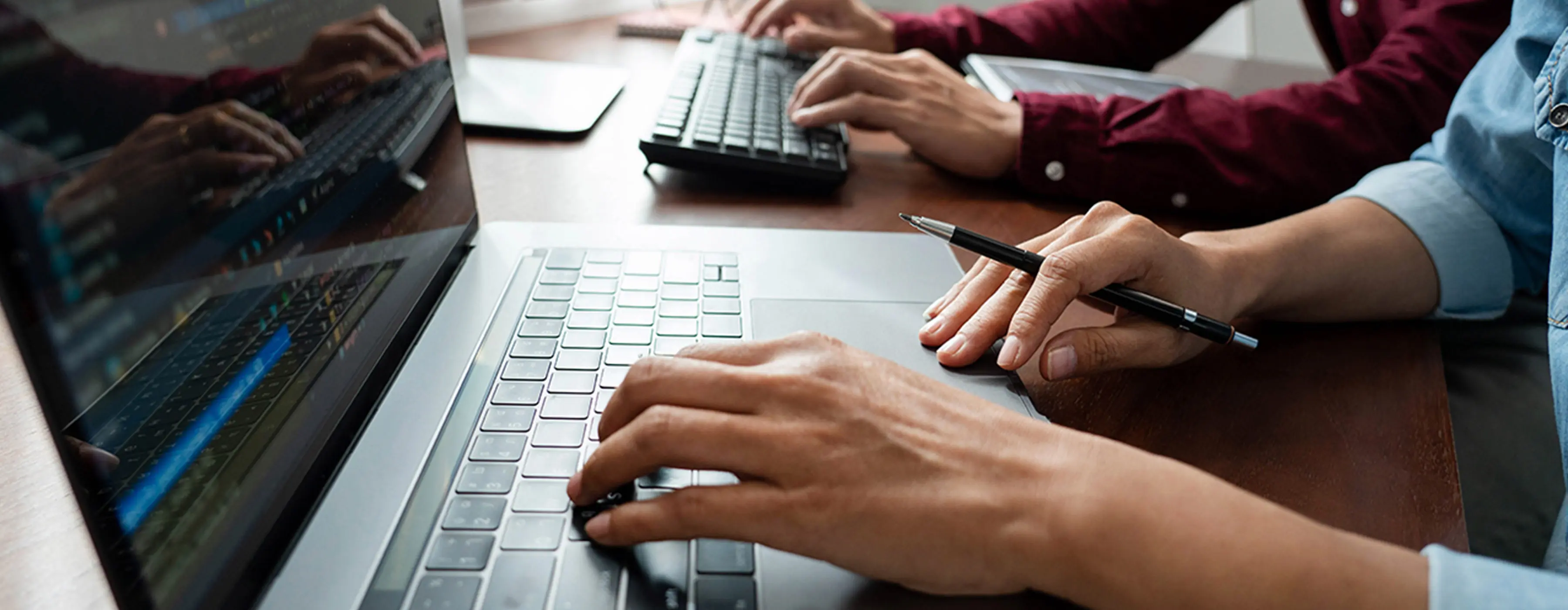 Foto em close de mãos de duas pessoas, uma teclando em notebook e outra em teclado em computador.
