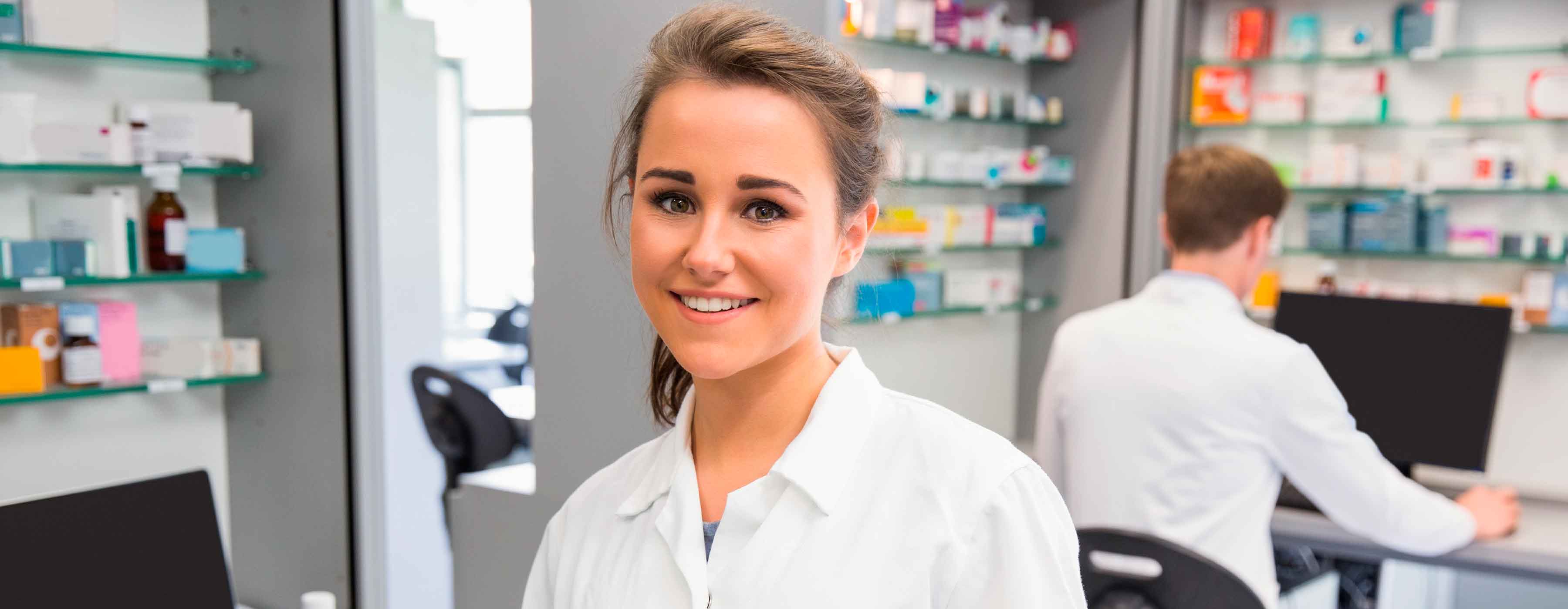 Mulher de jaleco em laboratório farmacêutico, com prateleiras de medicamentos ao fundo.