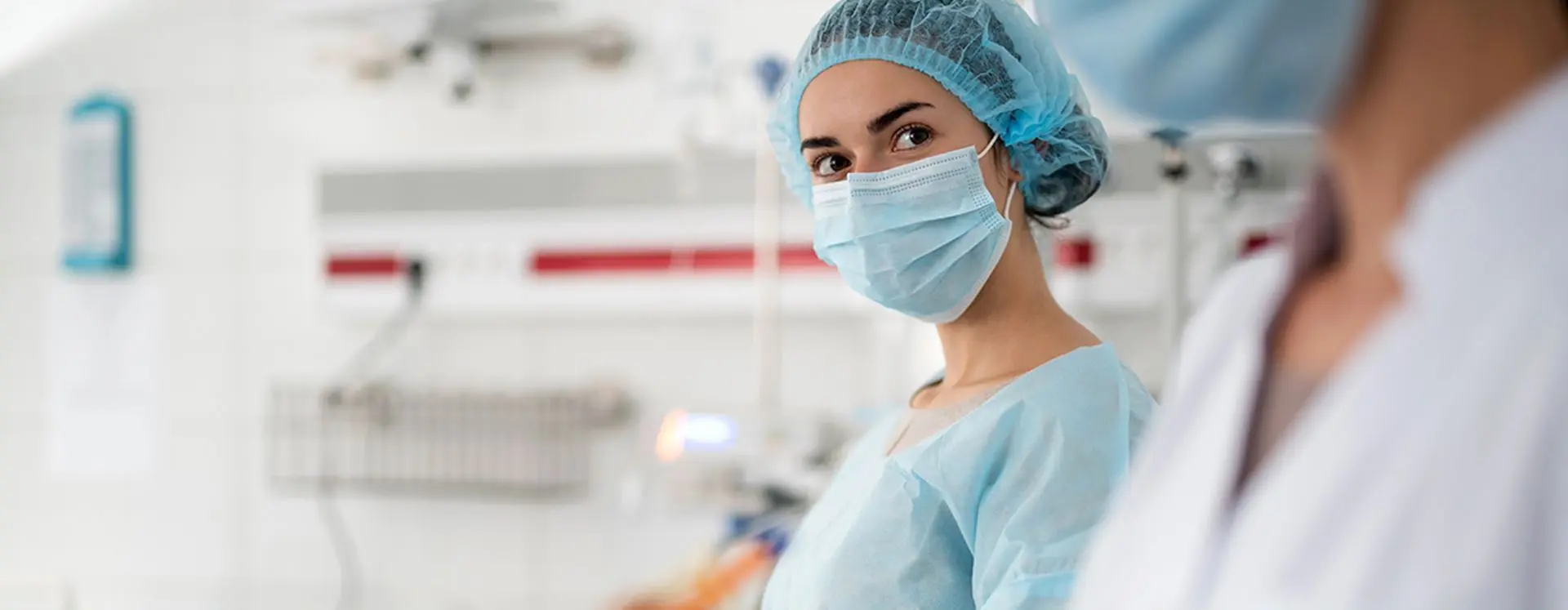 Foto de profissional da saúde uniformizada e usando máscara anotando dados em prancheta na frente de monitores de batimentos cardíacos e soros, ao fundo paciente entubado na cama hospitalar.