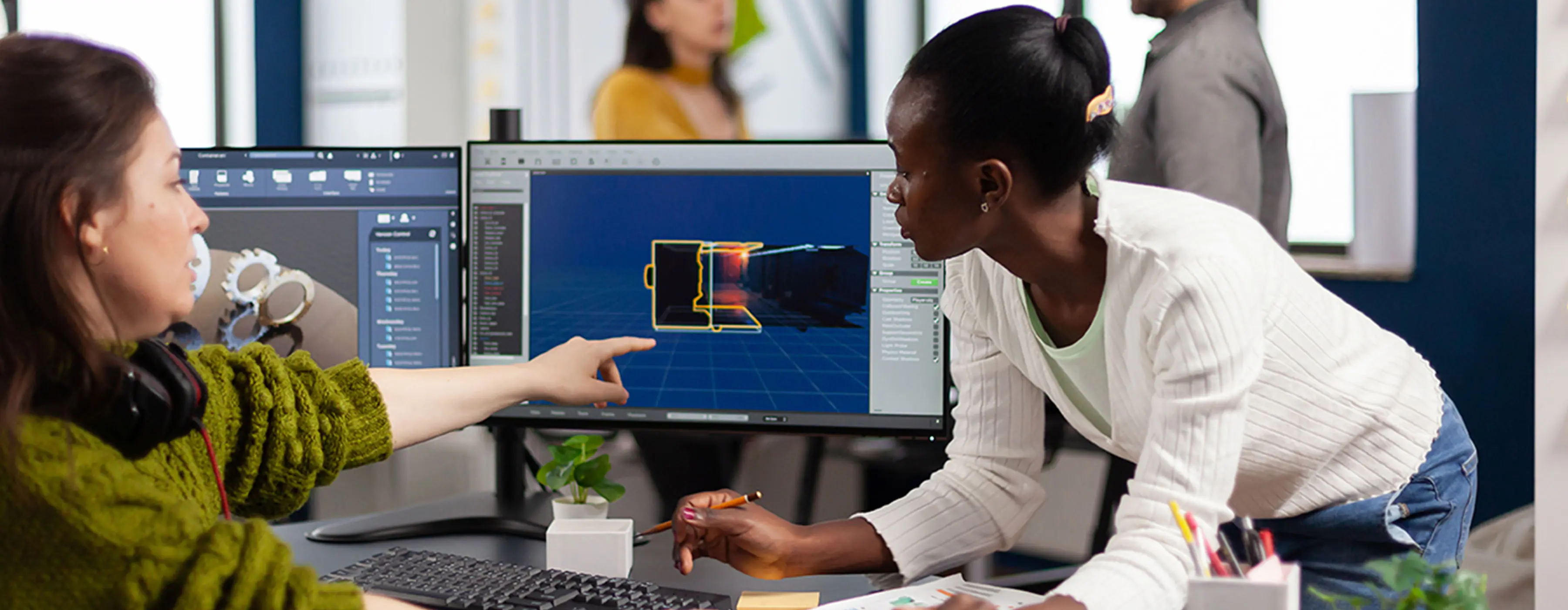 Foto de um mulher editando vídeo de game em computador com dois monitores e outra mulher observando as telas em escritório.