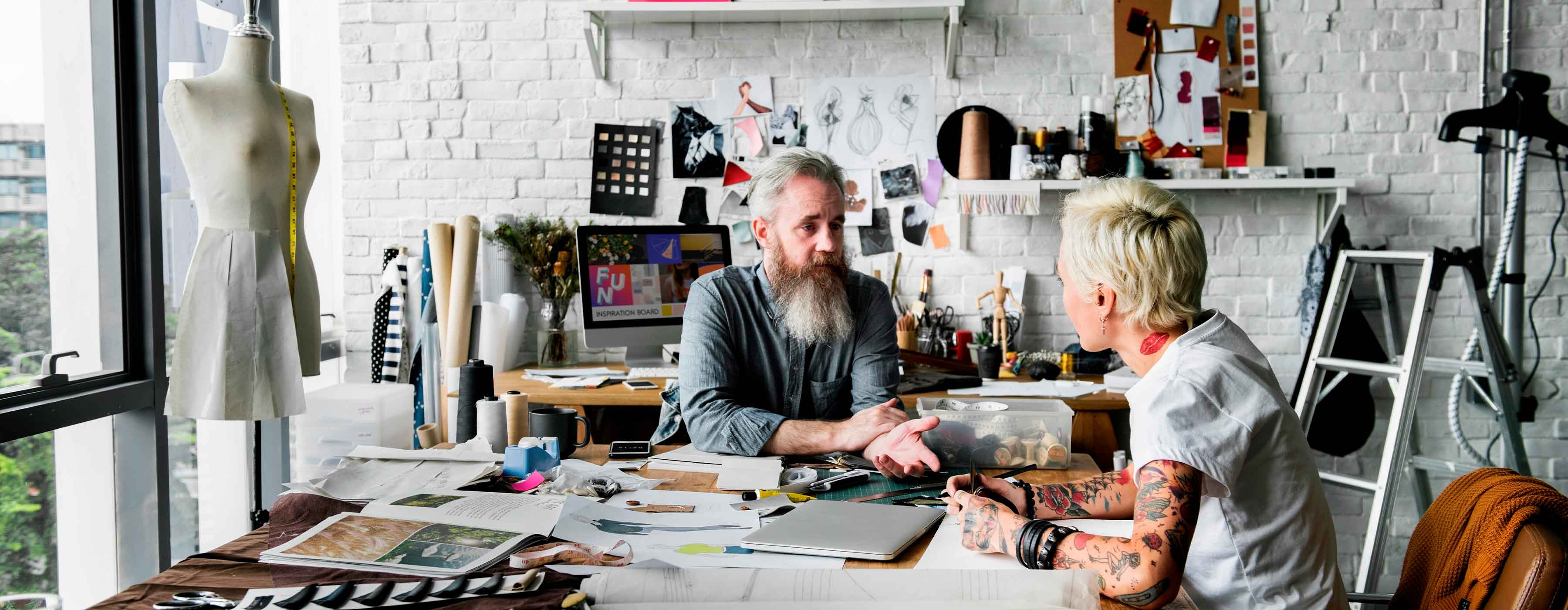 Pós-graduação em Gestão Estratégica em Negócios da Moda - foto de um rapaz com barba e uma moça, sentados, conversando em um ateliê de moda