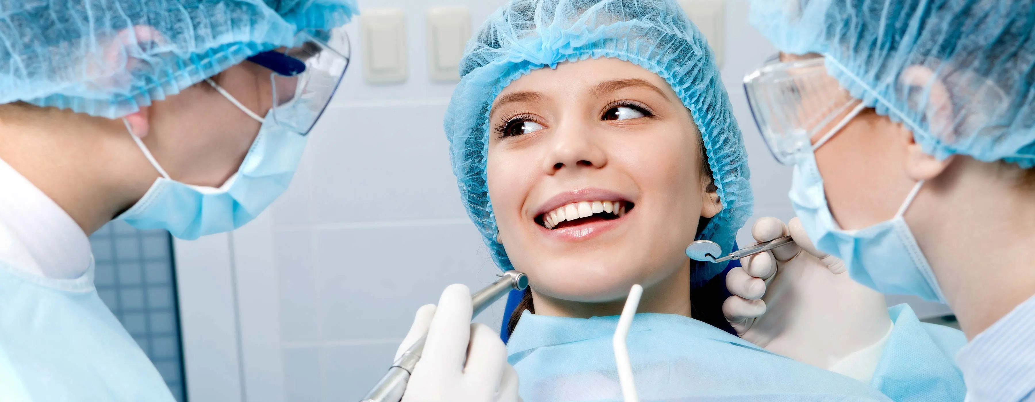 Foto de close de uma jovem sorrindo em cadeira odontológica e dois profissionais da saúde observando sua boca, um segura espelho e outro caneta de alta rotação em consultório odontológico.