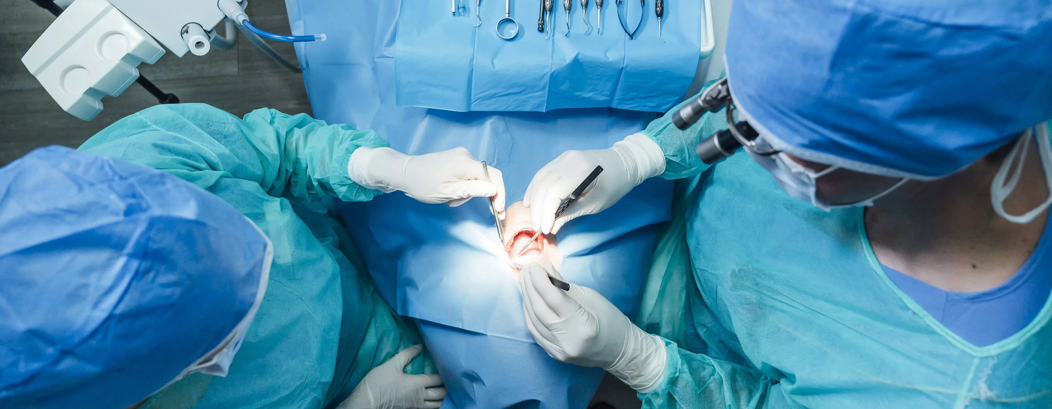 Foto com foco na boca de paciente em cadeira odontológica, com dois profissionais de saúde bucal manuseando com pinças cirúrgicas em consultório odontológico.