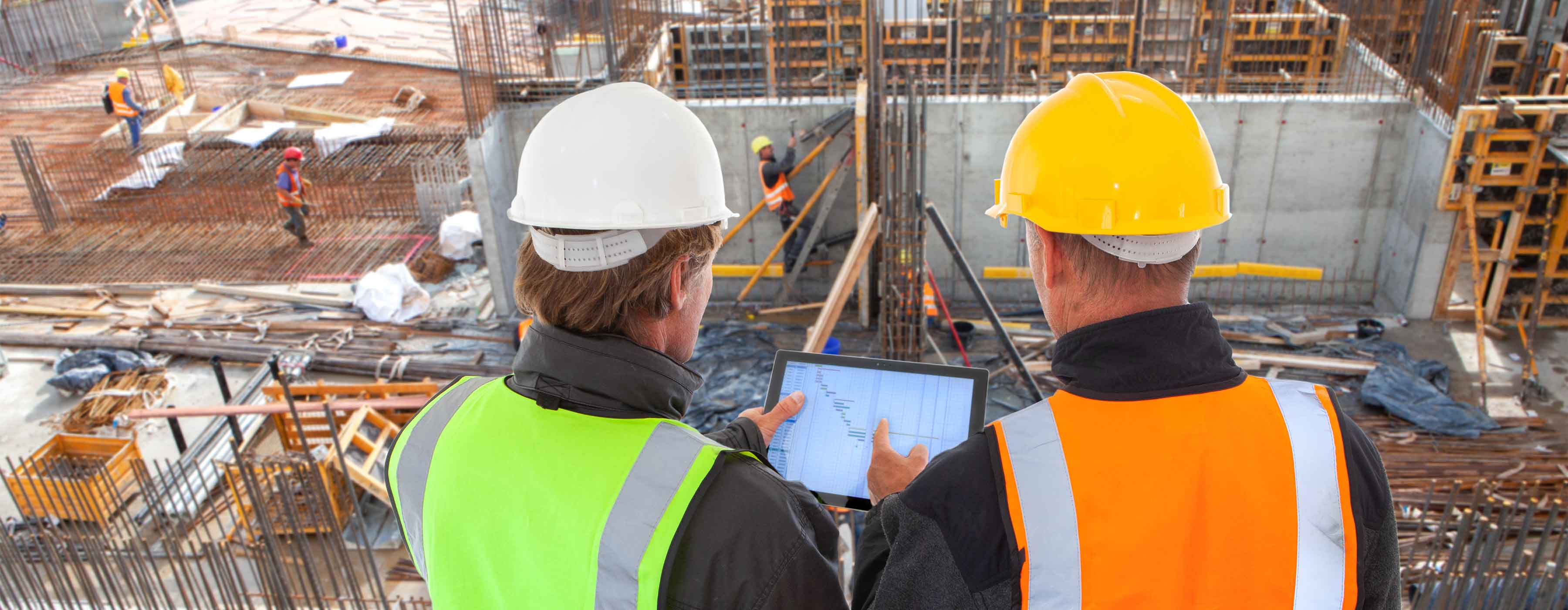 Dois profissionais com capacetes e coletes de segurança analisam planta em tablet em obra com trabalhadores ao fundo.