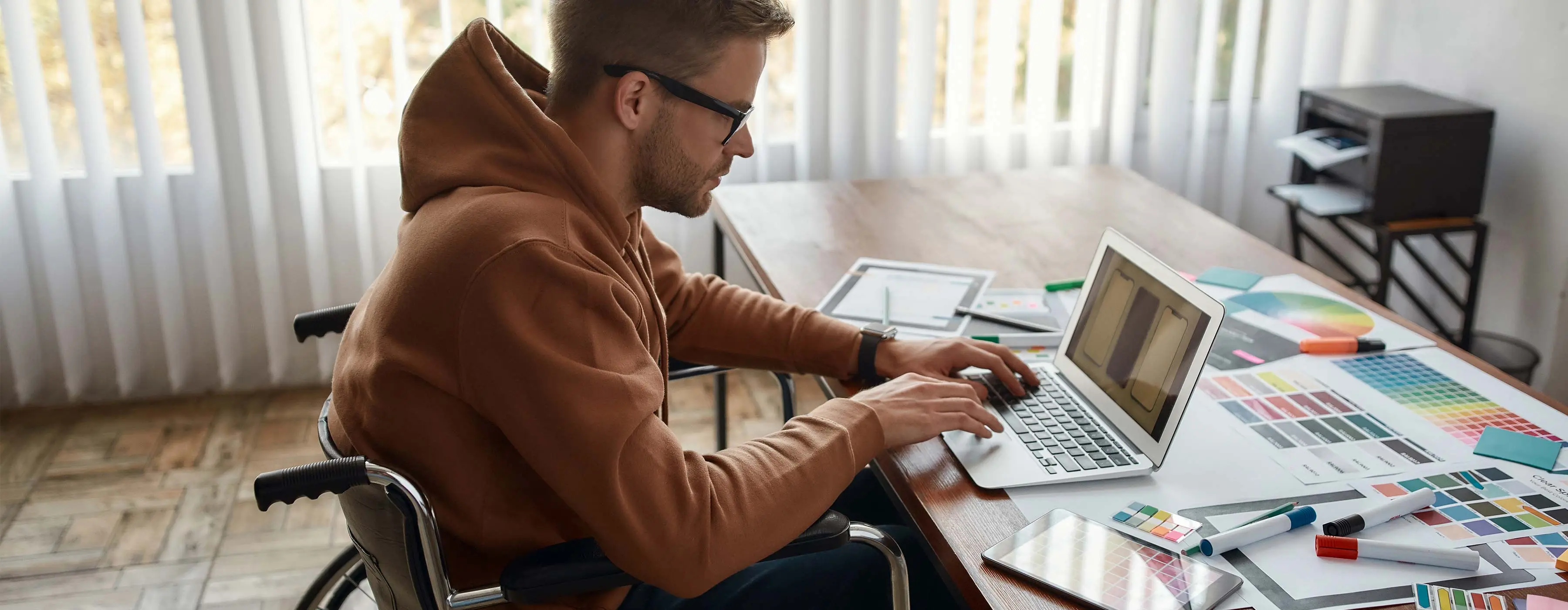 Foto de homem em cadeiras de rodas em frente a uma mesa usando notebook em meio a papéis com diversas cores