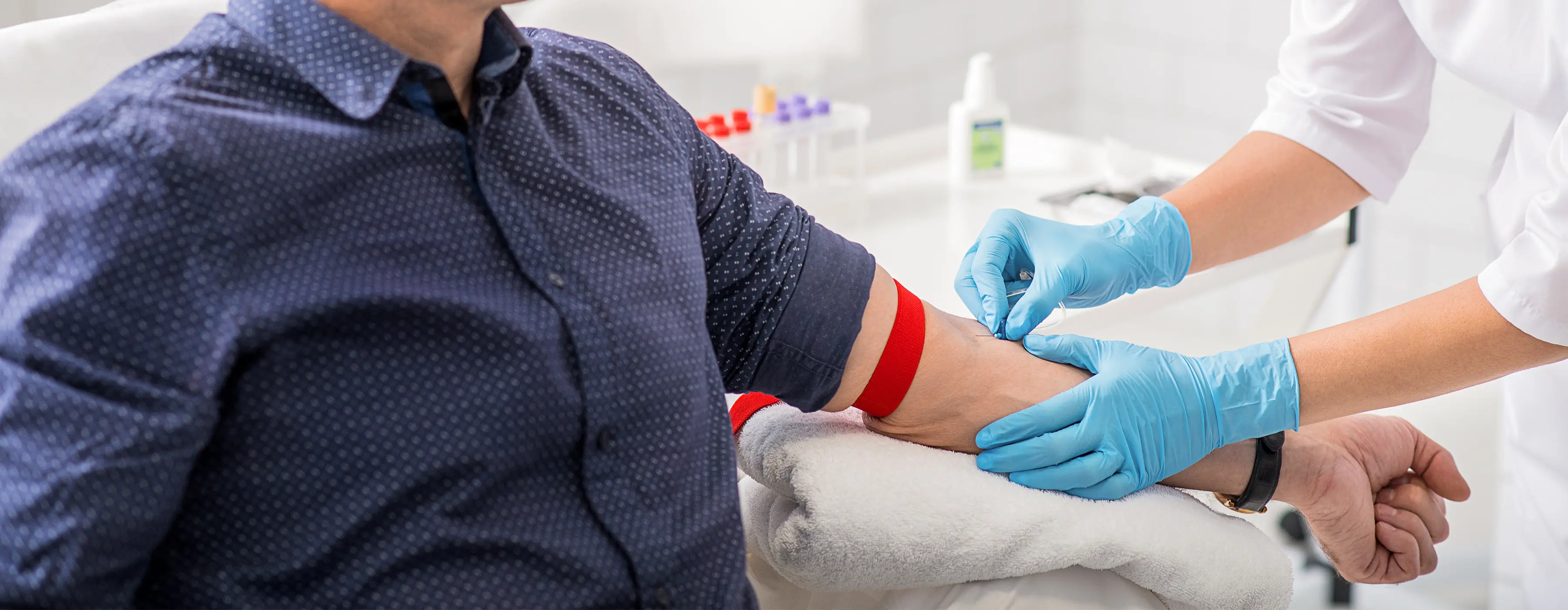 Pessoa em laboratório vendo amostra de sangue por um microscópio. Ao seu lado, há vários tubos de sangue.