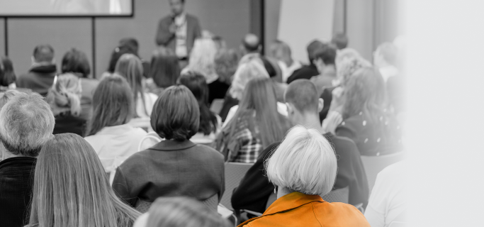Plateia de costas assistindo a uma palestra em auditório, com destaque para uma pessoa em primeiro plano usando jaqueta laranja. Ao fundo, um palestrante de pé com microfone. Imagem em preto e branco.