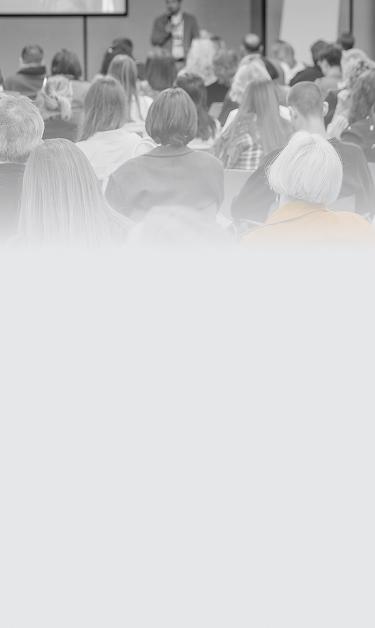 Plateia de costas assistindo a uma palestra em auditório, com destaque para uma pessoa em primeiro plano usando jaqueta laranja. Ao fundo, um palestrante de pé com microfone. Imagem em preto e branco.