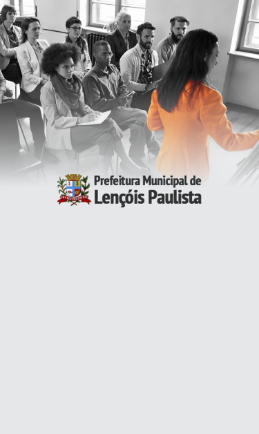 Foto em preto e branco de grupo de pessoas sentadas, uma mulher de casaco laranja fala e aponta para uma lousa.