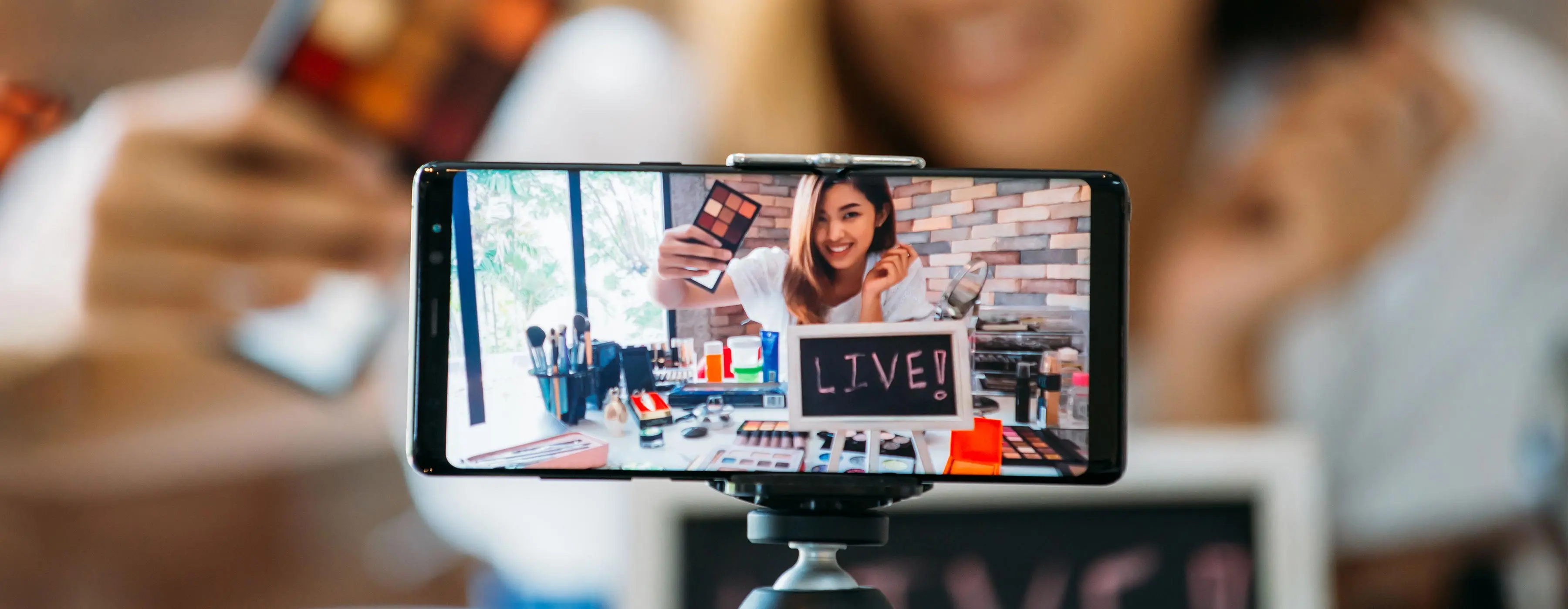 Foto em close de um celular onde aparece uma mulher gravando vídeo com produtos de maquiagem.