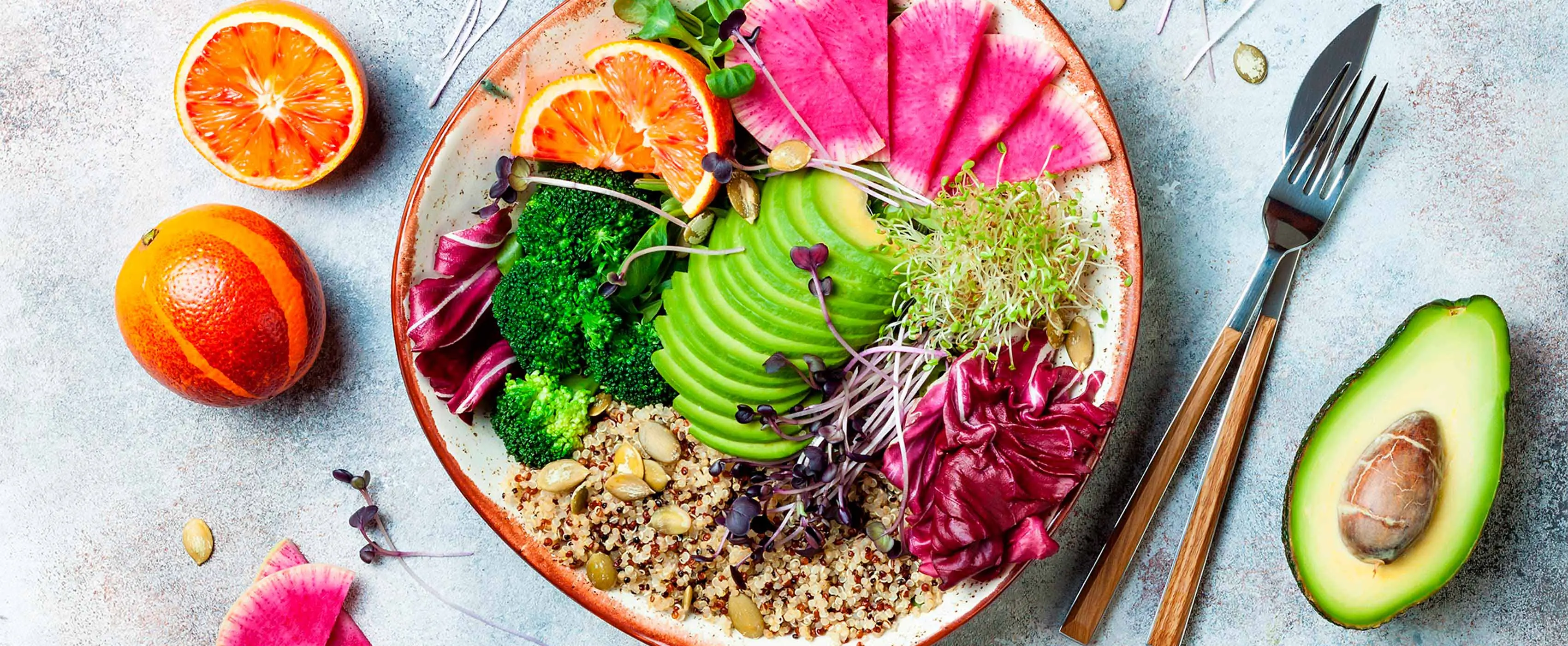 Foto em close de um prato repleto de verduras e frutas, acompanhado por talheres e mais frutas ao redor.