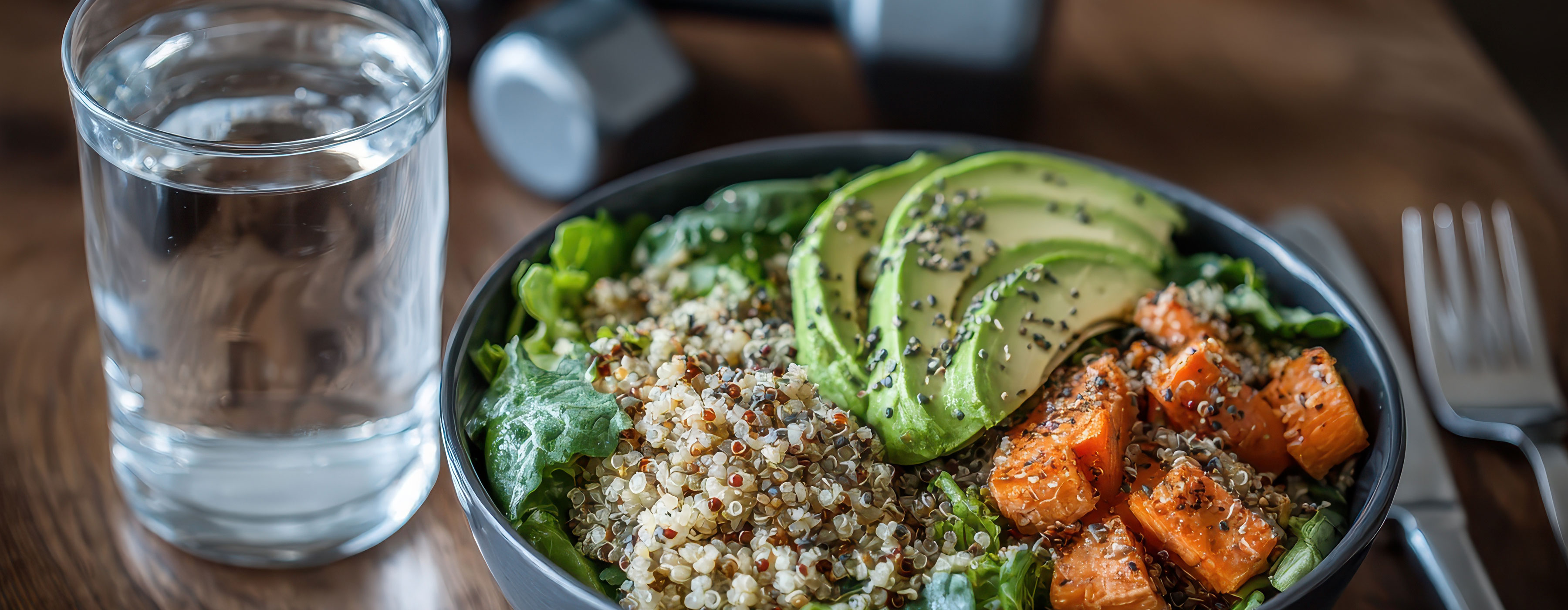 Nutrição Aplicada ao Exercício Físico - foto de uma tigela com salada saudável de folhas verdes, legume, abacate,  sementes ao lado de um  copo de água, talher e alteres de ginástica ao fundo.