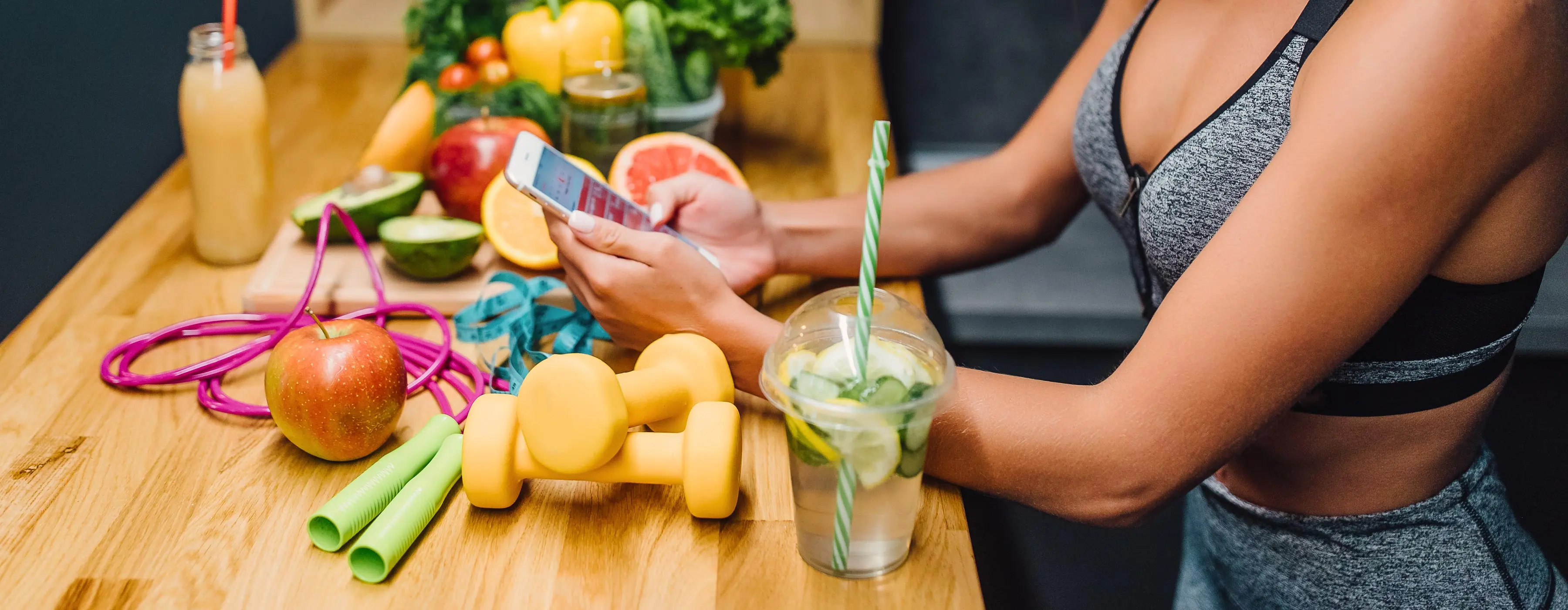 Foto de uma mulher com roupa de ginástica usando celular e comendo frutas e água aromatizada.