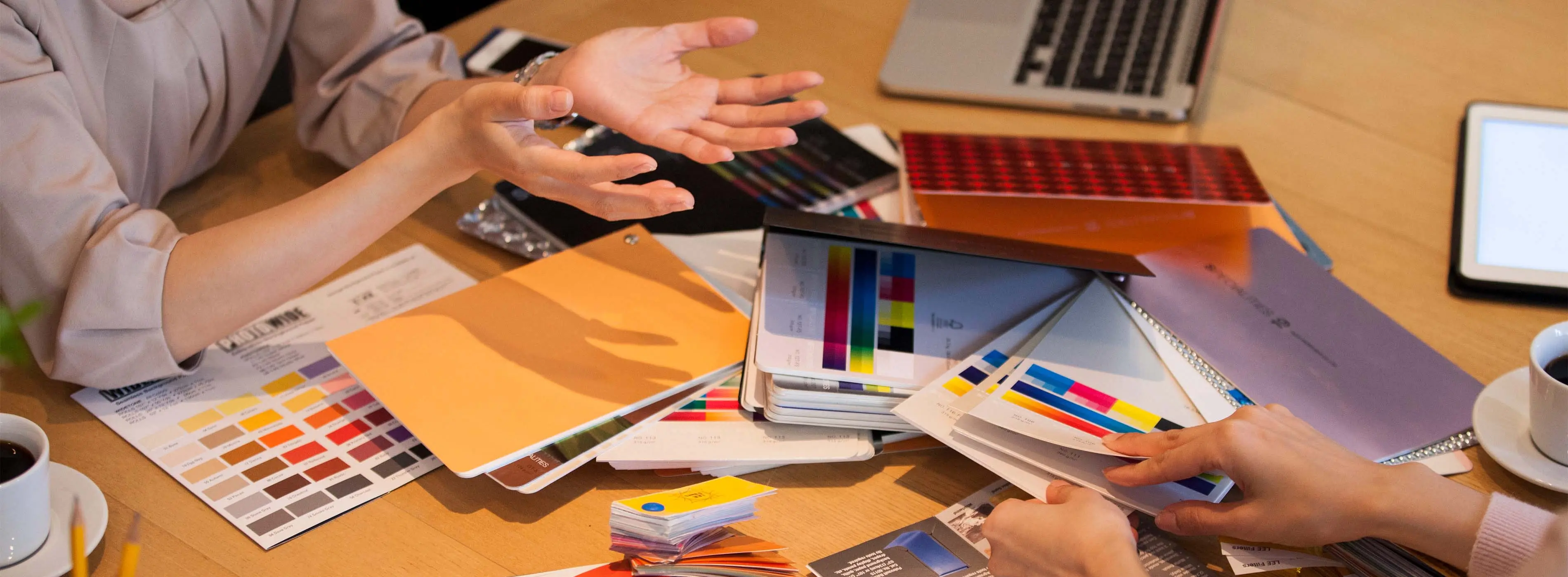 Foto focando nas mãos de duas pessoas em reunião com cartela de cores e notebook, cadernos e tablet em mesa de escritório.