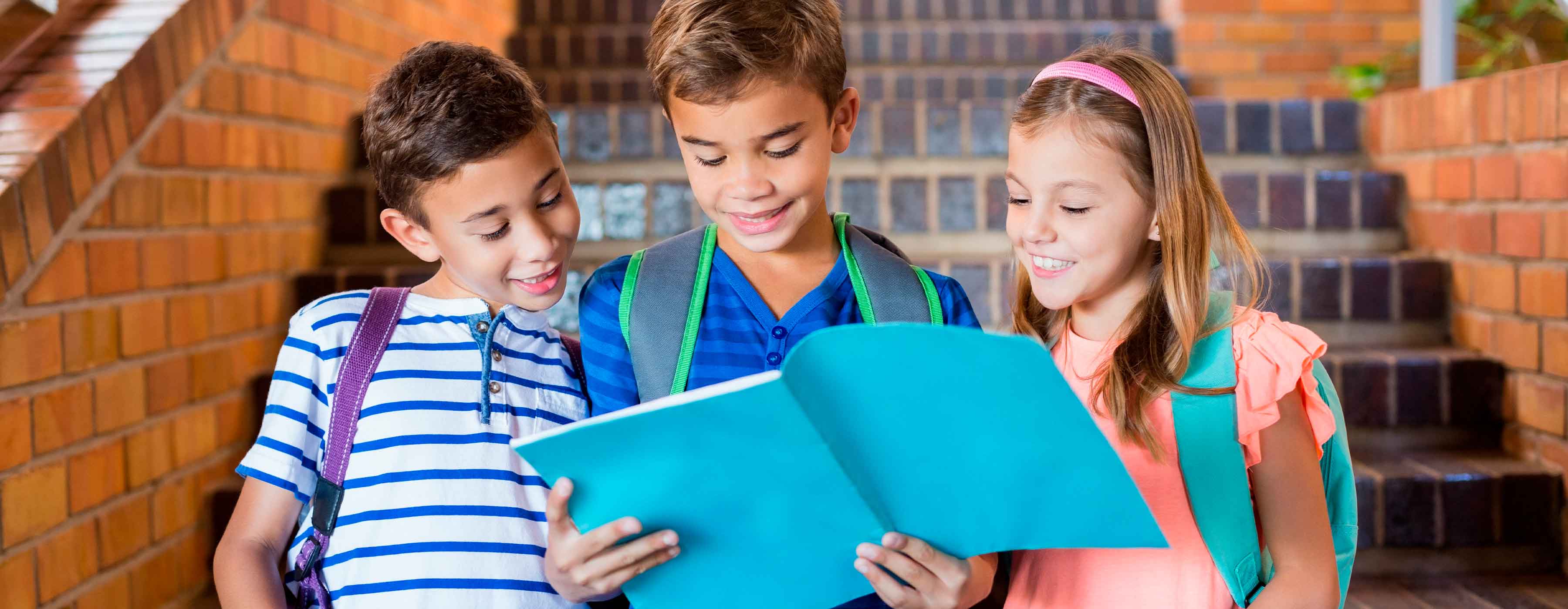 Inglês ¿ Junior 2 ¿ foto de três crianças com mochilas escolares lendo juntas um livro de capa azul.