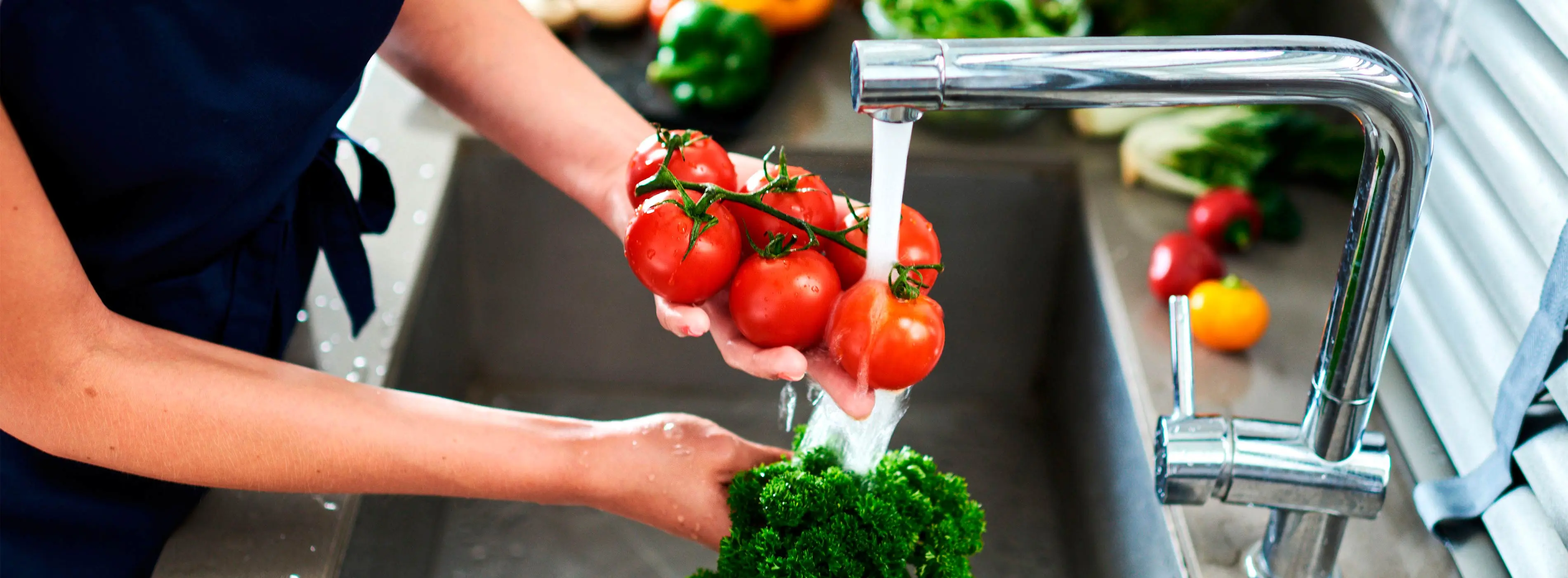 Foto em close de mãos de uma pessoa segurando punhado de tomates rasteiro lavando na pia de uma cozinha.