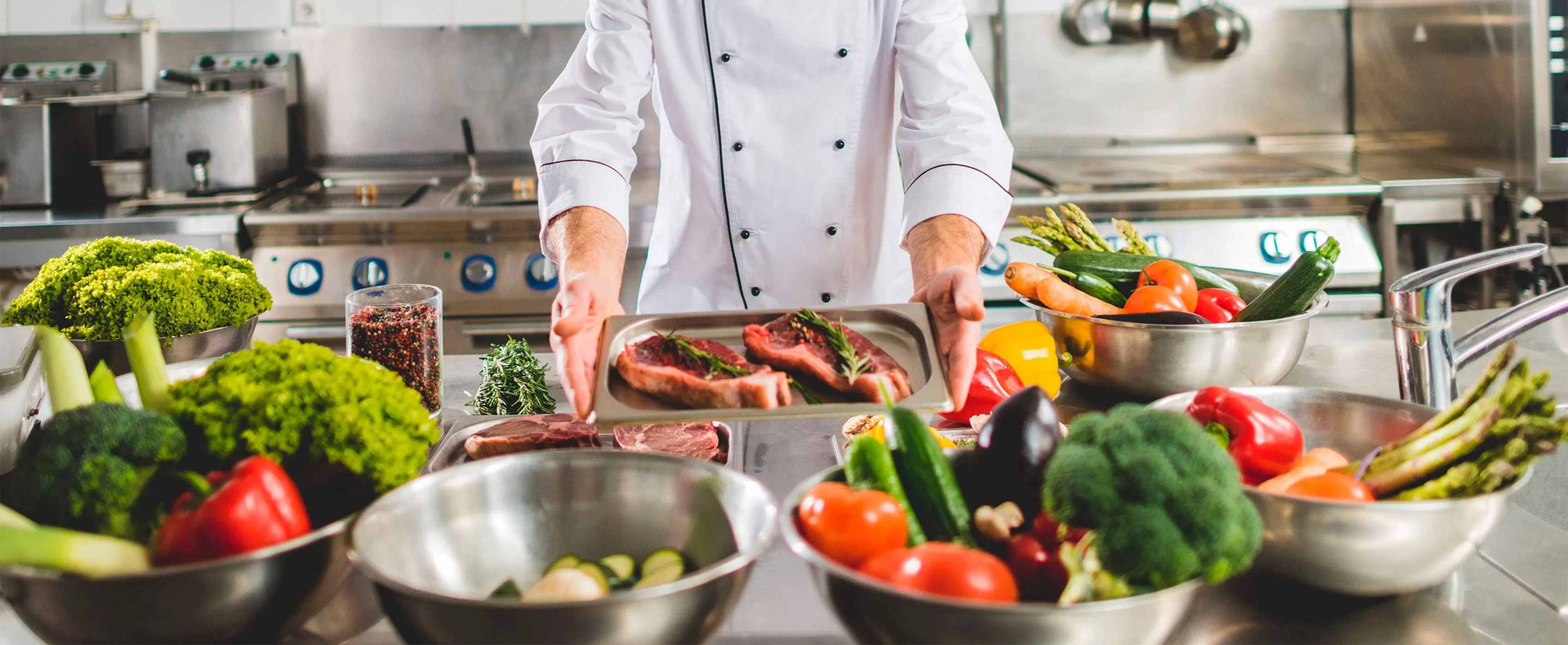 Foto em close de mãos de chef mostrando carne em bandeja temperada crua em mesa com tigelas com legumes em cozinha profissional.