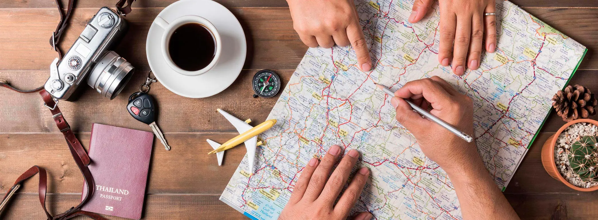 Foto em close das mãos de duas pessoas verificando um mapa em cima da mesa. Ao lado do mapa, uma miniatura de avião, bússola, passaporte, uma chave e uma xícara de café.
