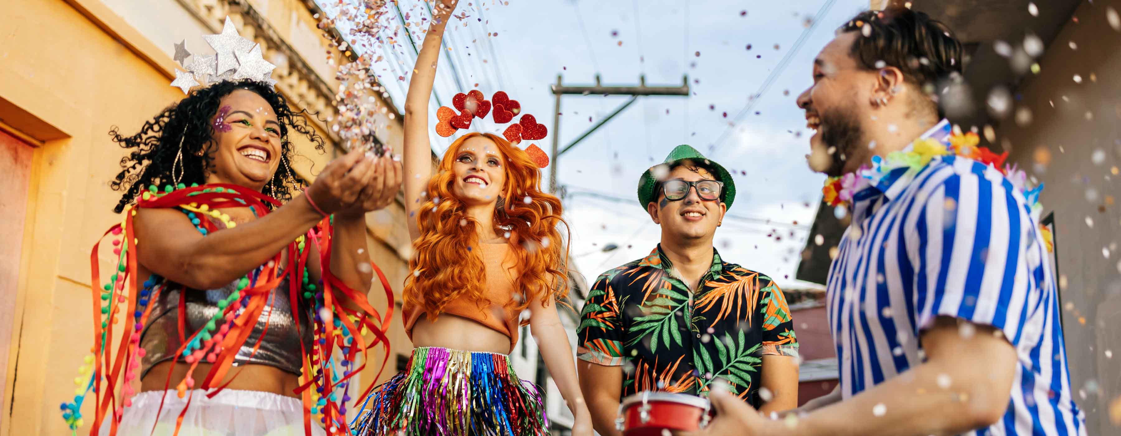 Foto de grupo de pessoas em ambiente externo festivo, usando roupas coloridas e acessórios, enquanto jogam confetes para o alto.