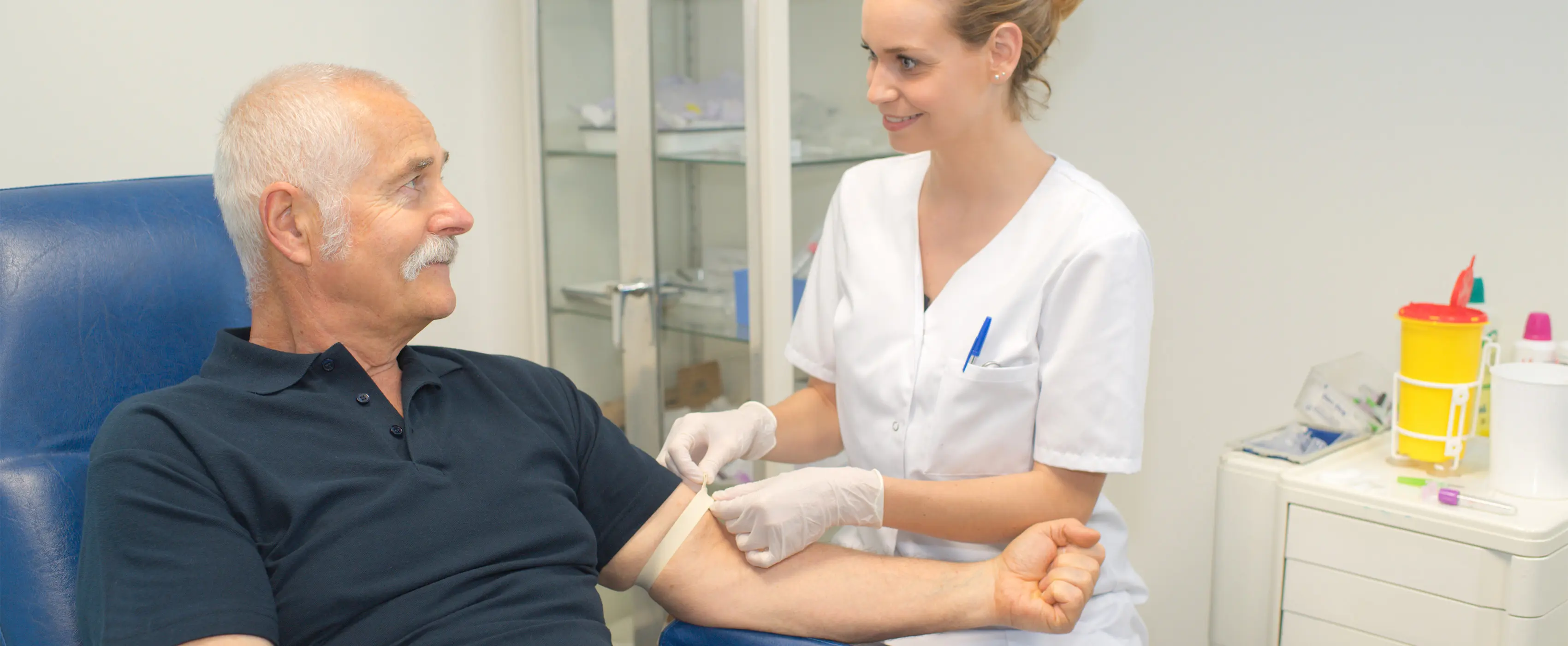 Foto de um homem sendo preparado por uma mulher com roupa de enfermeira para fazer a coleta de sangue em um laboratório clínico.