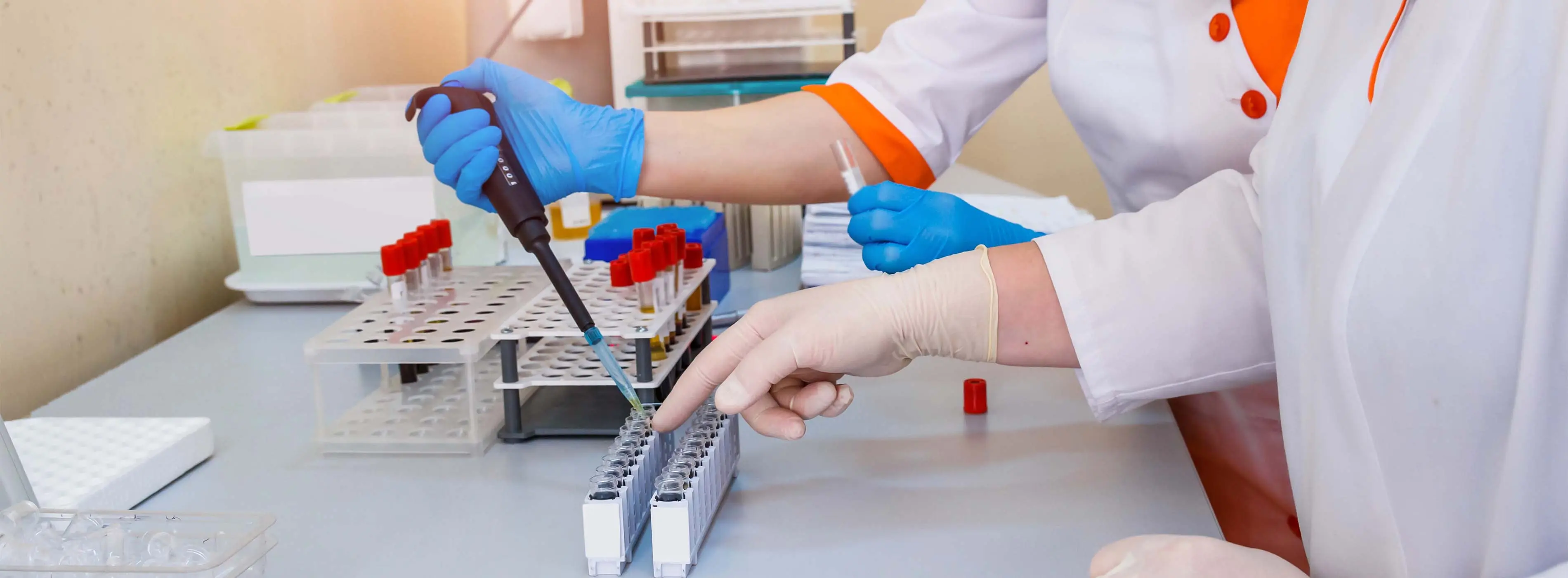 Foto de duas pessoas com jaleco e luvas cirúrgicas manipulando materiais sanguíneos com ferramentas de enfermagem em um cenário de laboratório clínico.