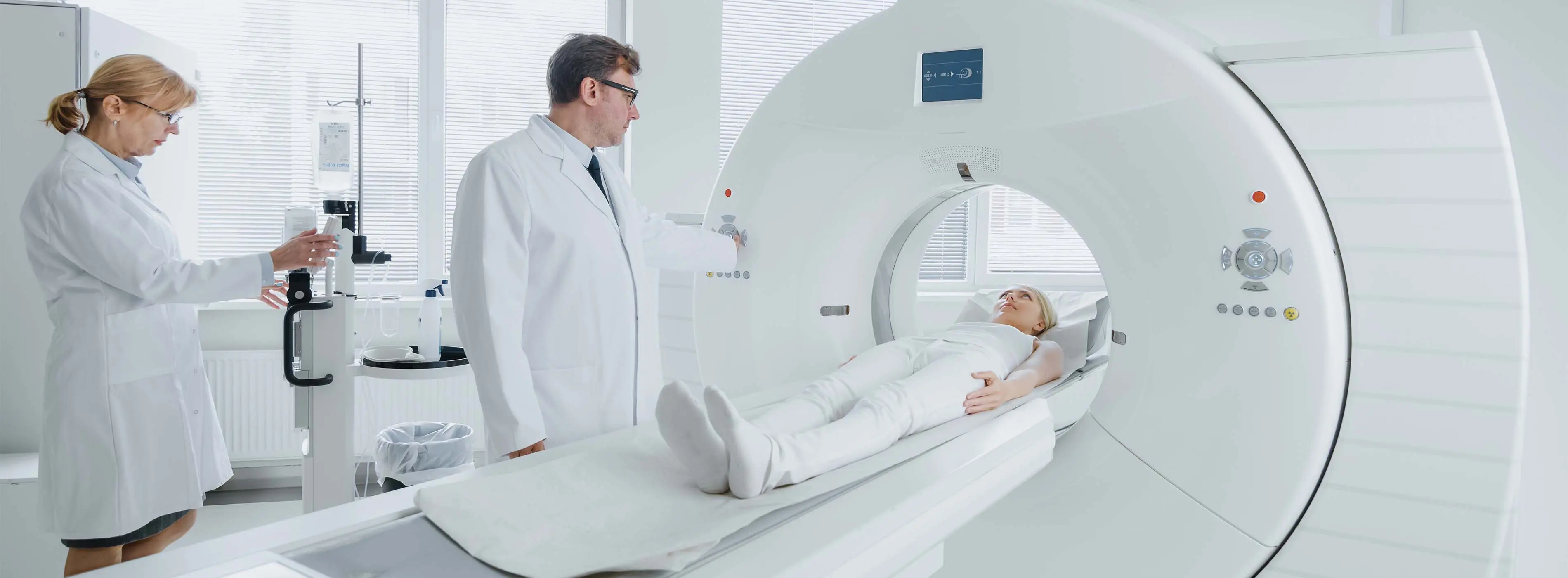 Foto de mulher deitada em uma máquina de tomografia que está sendo manipulada por duas pessoas vestidas com jalecos brancos.