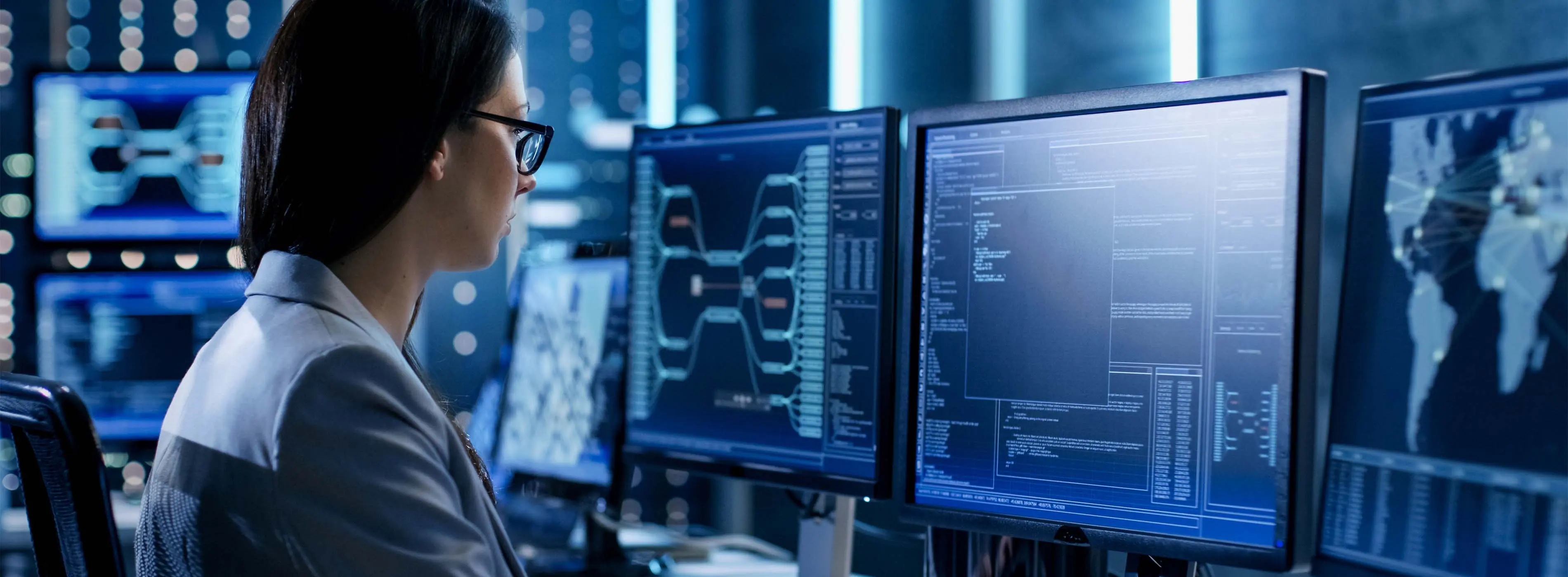 Foto de mulher observando códigos em três telas de monitores em sala de computadores.