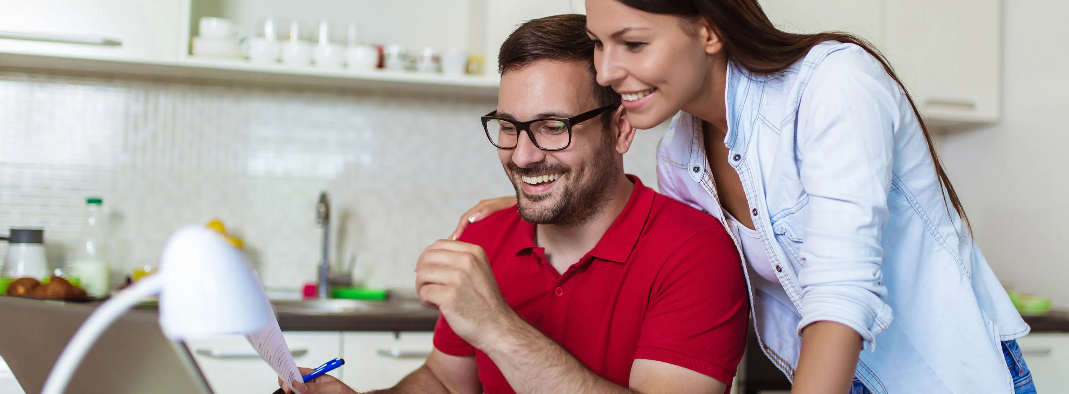 Foto de um casal checando papel e notebook em cozinha de residência.