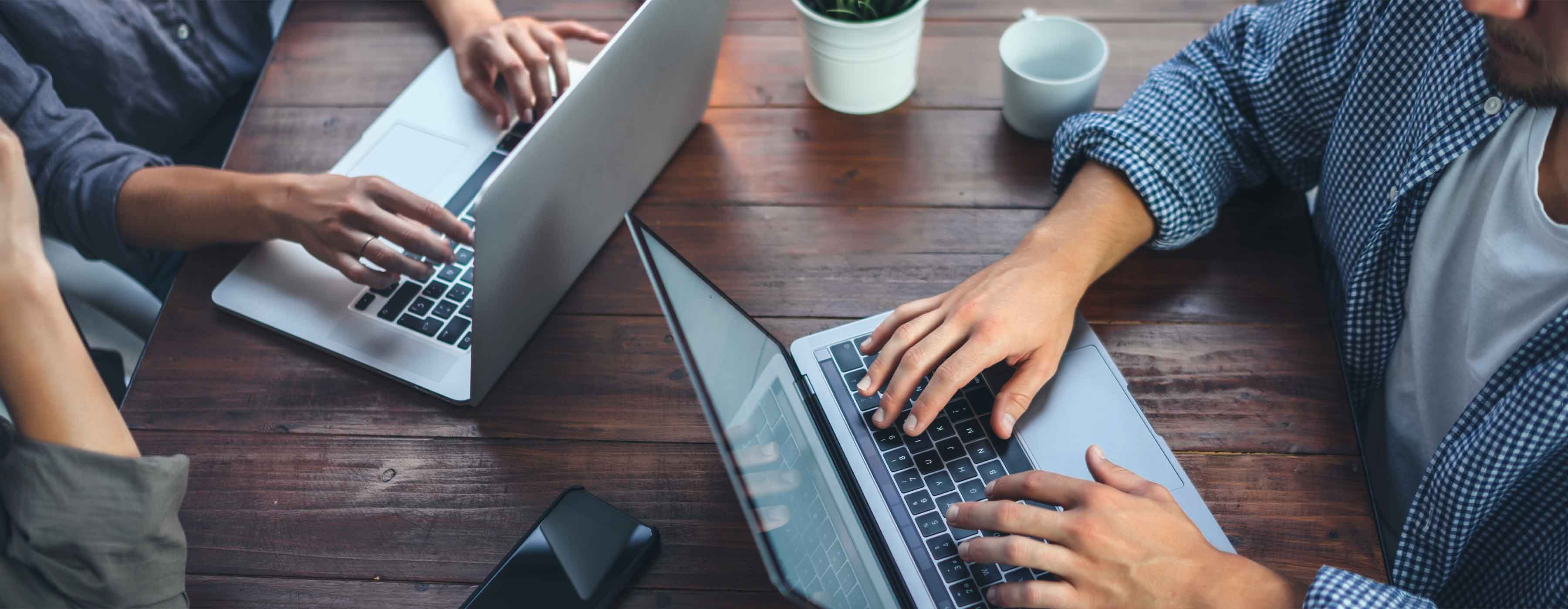 Duas pessoas usando notebooks sobre uma mesa de madeira, com as mãos digitando nos teclados.